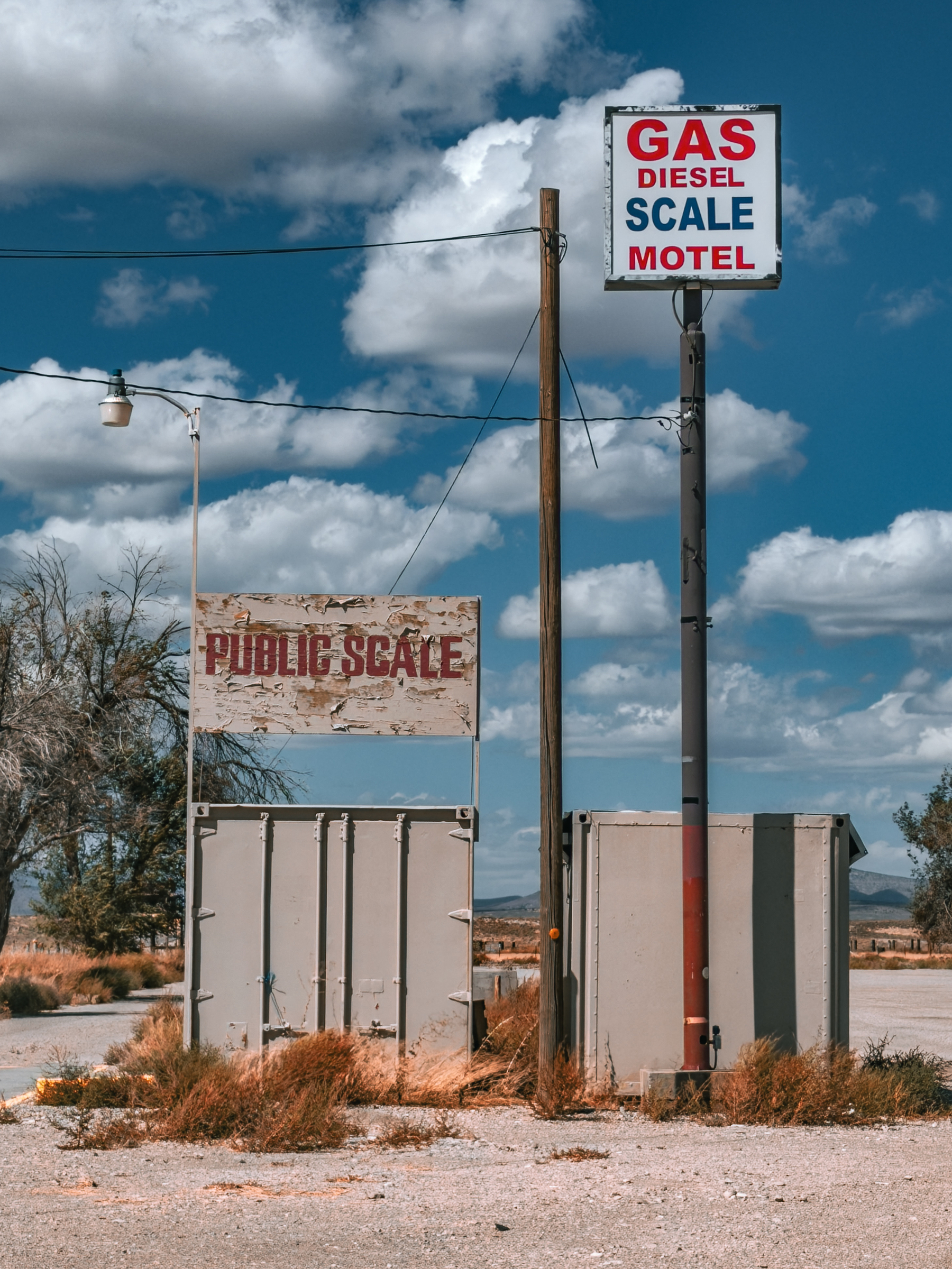 Auto-generated description: A roadside setup displays signs for gas, diesel, scale, motel, and a weathered public scale in a rural area under a partly cloudy sky.