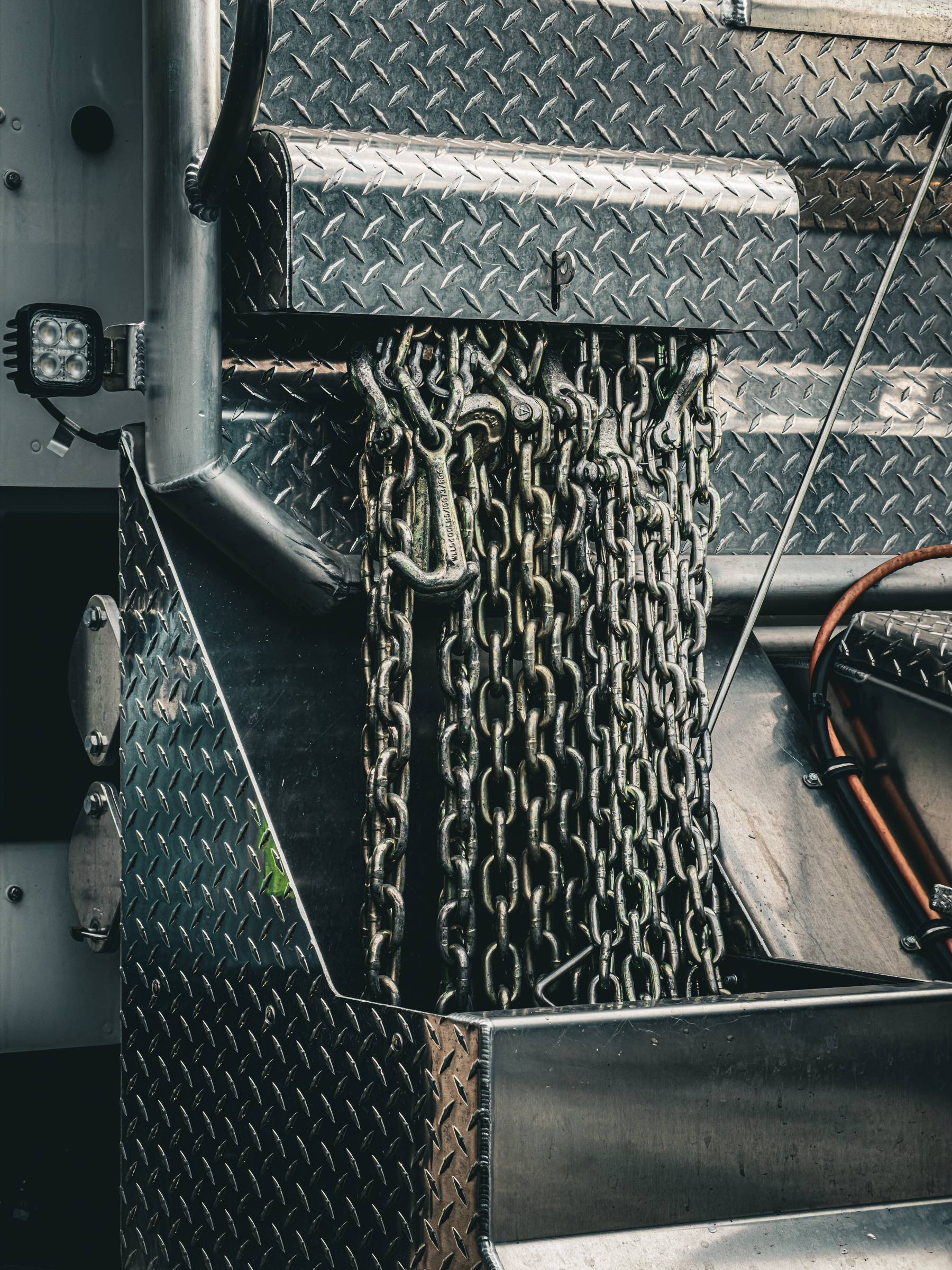 Auto-generated description: Heavy metal chains with hooks are hanging from a compartment on a diamond plate metal surface.