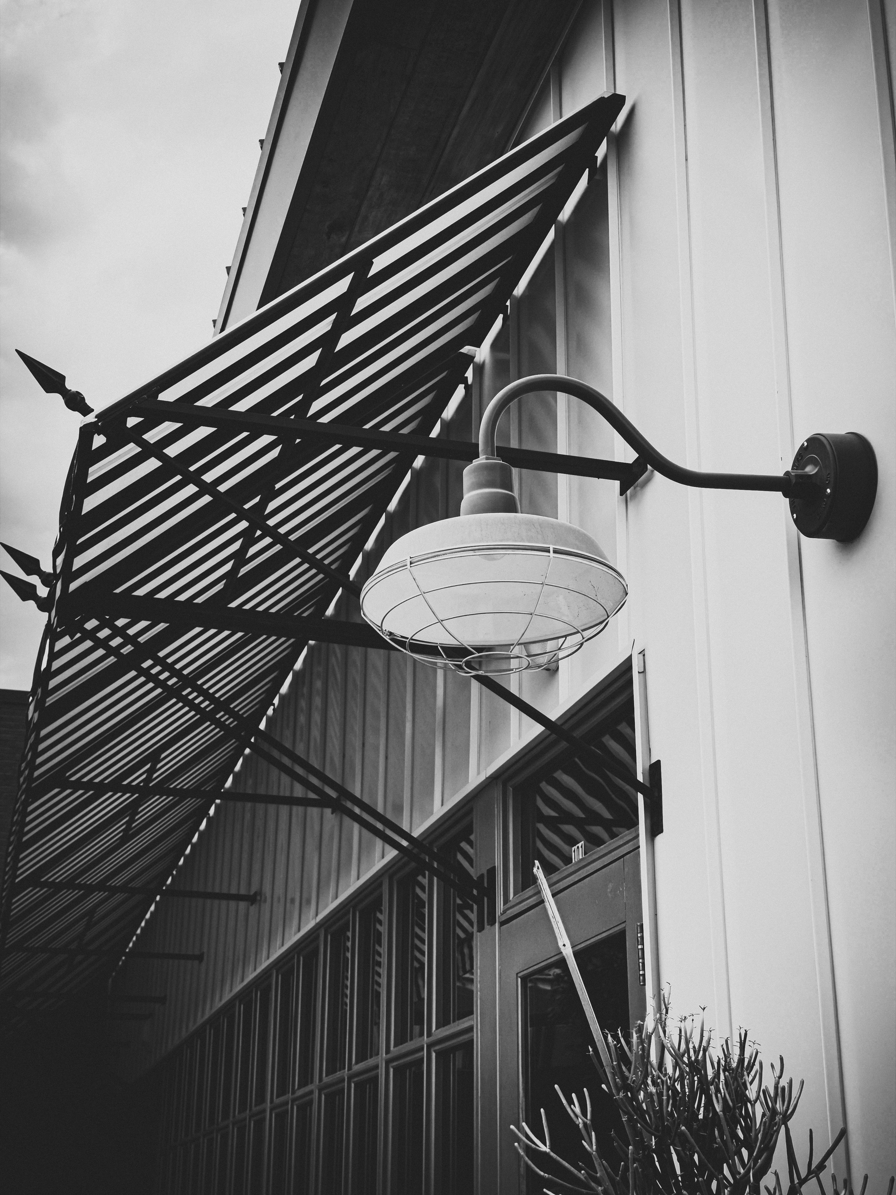 Auto-generated description: A black and white photo showcases an exterior wall with a vintage industrial-style light fixture and a metal awning above a door and windows.