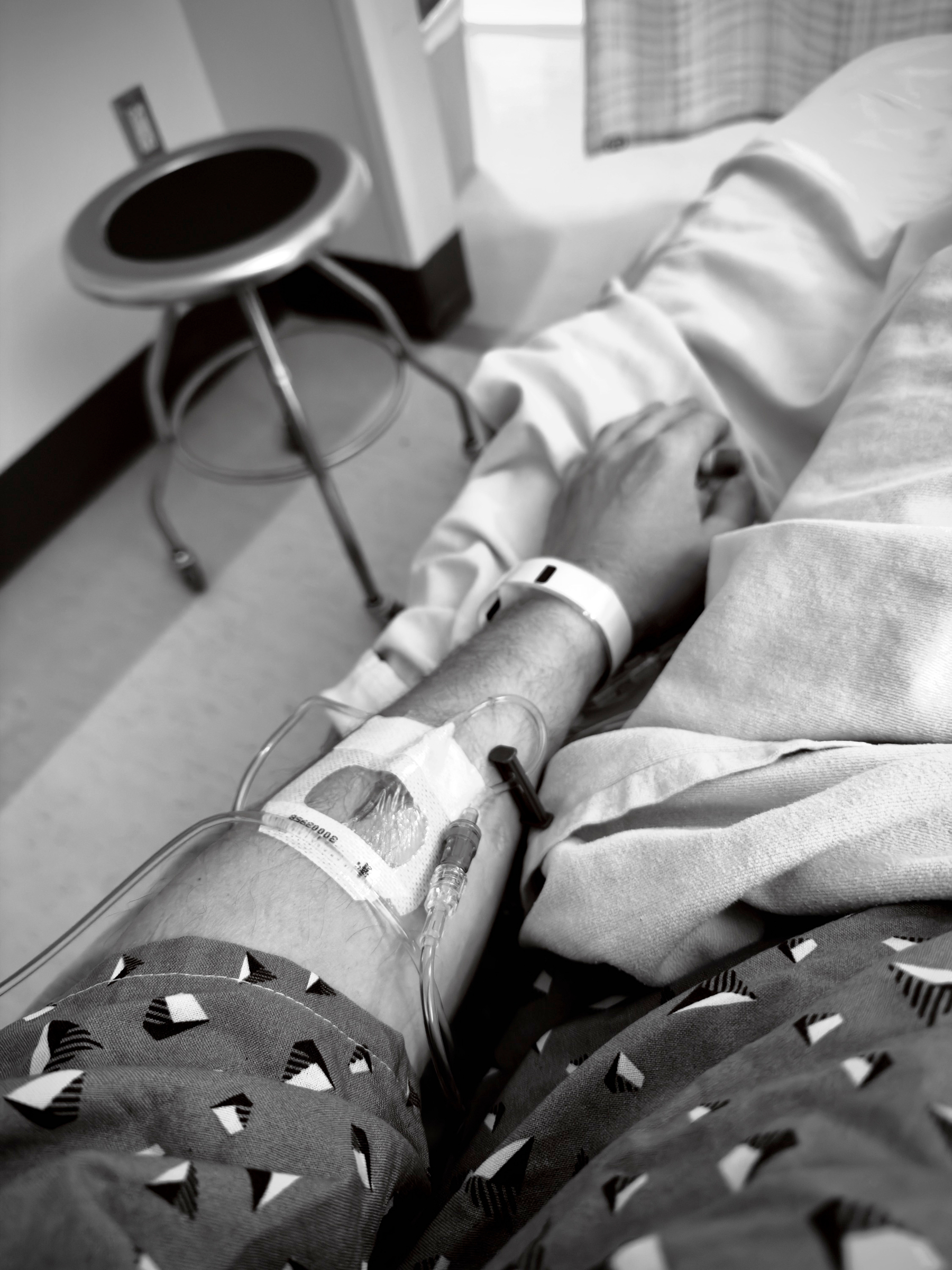 Auto-generated description: A person lying in a hospital bed with an IV inserted in their arm, wearing a hospital gown and an identification wristband.