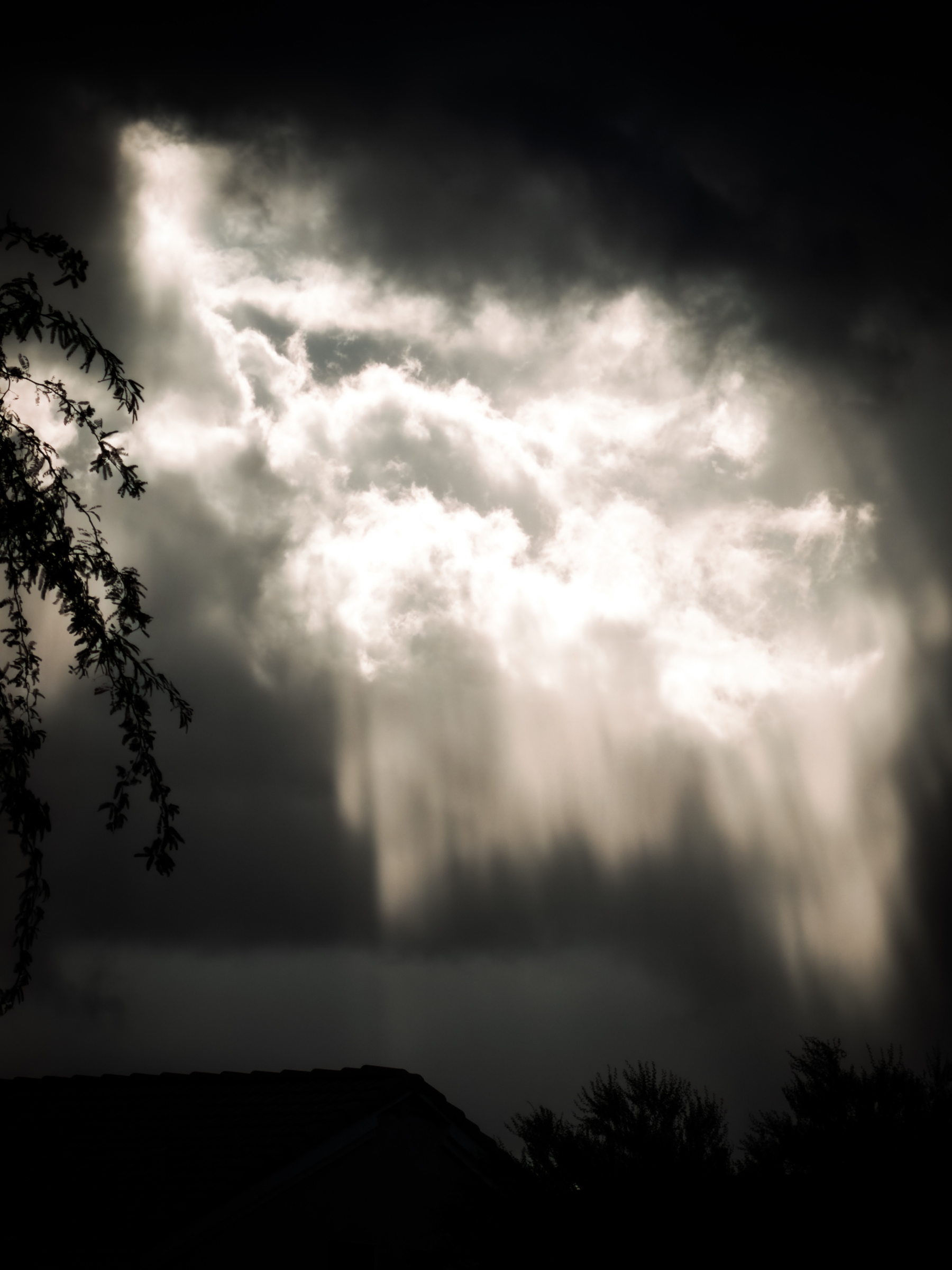 Auto-generated description: Sun rays break through dark, heavy clouds over silhouetted trees and rooftops.