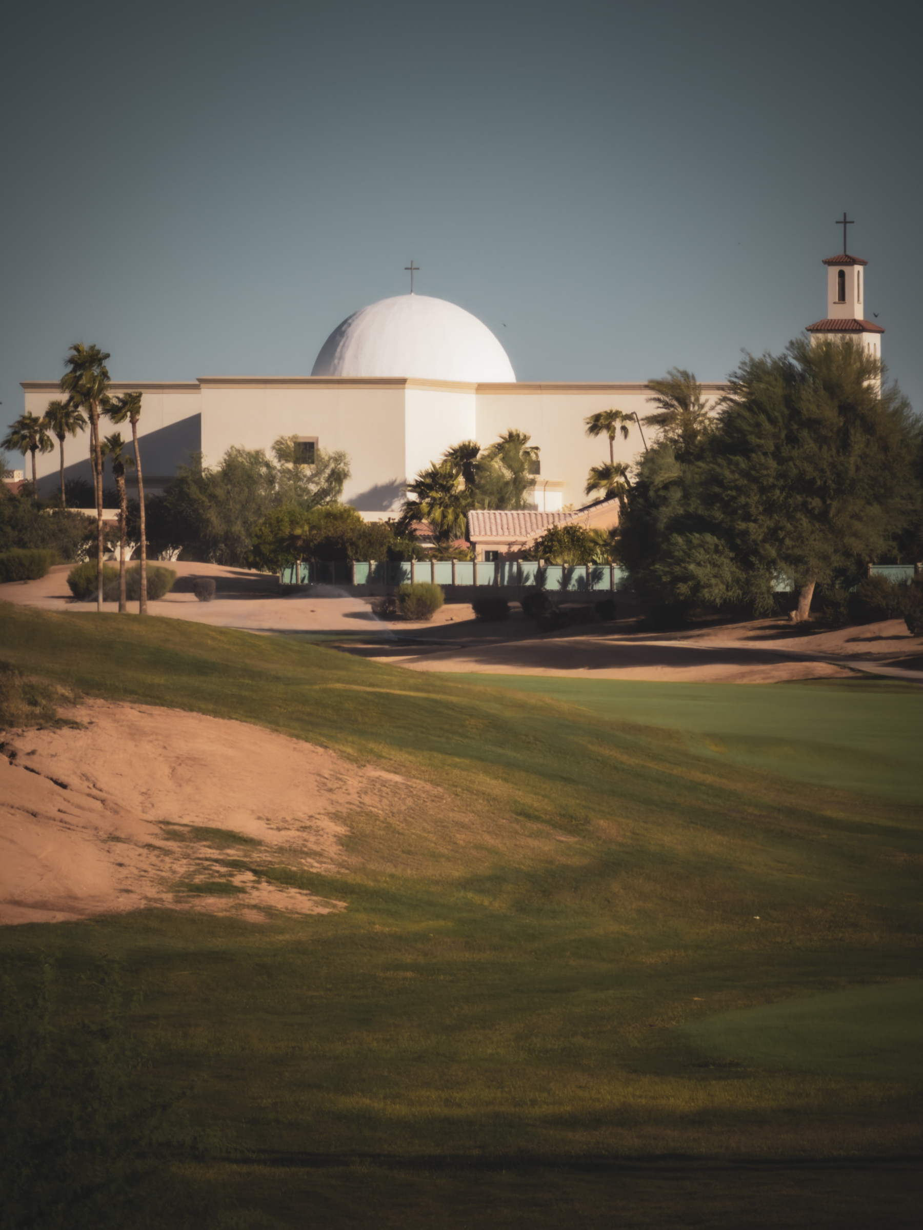 Auto-generated description: A white building with a dome and a tower topped with crosses stands behind a green golf course with scattered palm trees and shrubs.
