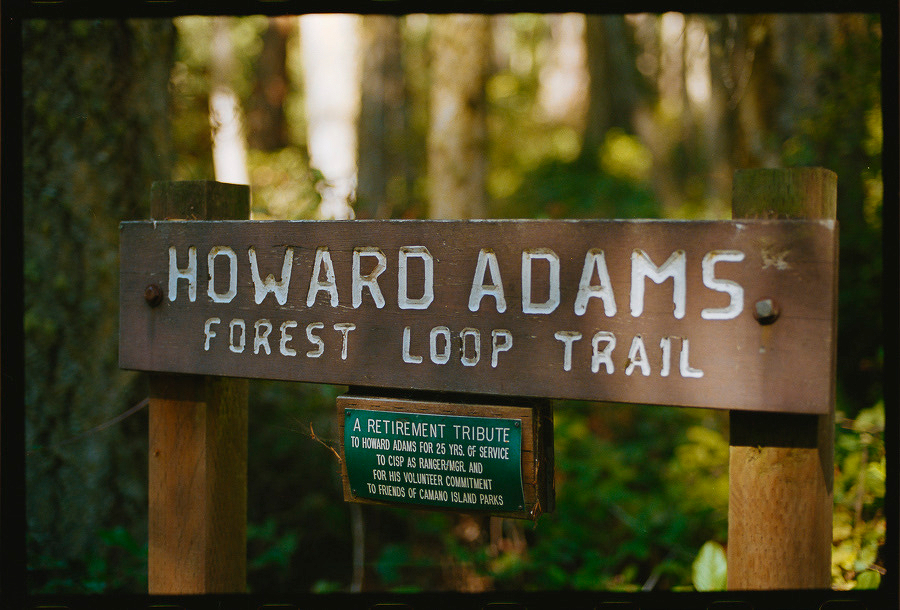Auto-generated description: A wooden sign marking the Howard Adams Forest Loop Trail stands in a wooded area, accompanied by a smaller plaque honoring Howard Adams for his service and volunteer commitment.