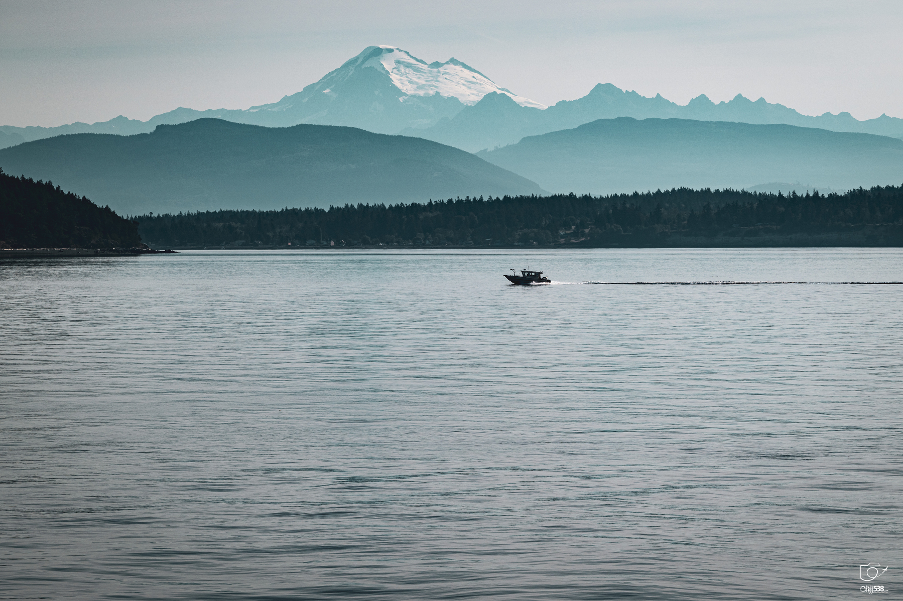 Auto-generated description: A boat is cruising across a calm body of water with a snow-capped mountain and forested hills in the background.