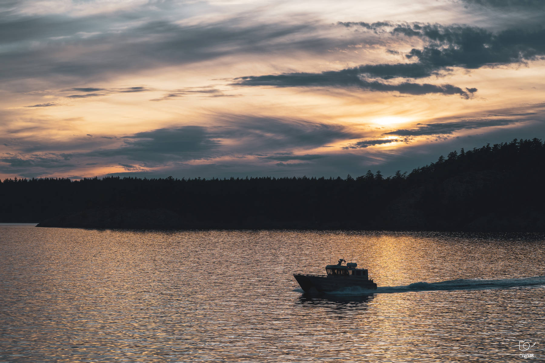 Auto-generated description: A boat is sailing across calm waters under a dramatic sunset sky with silhouetted trees on the horizon.