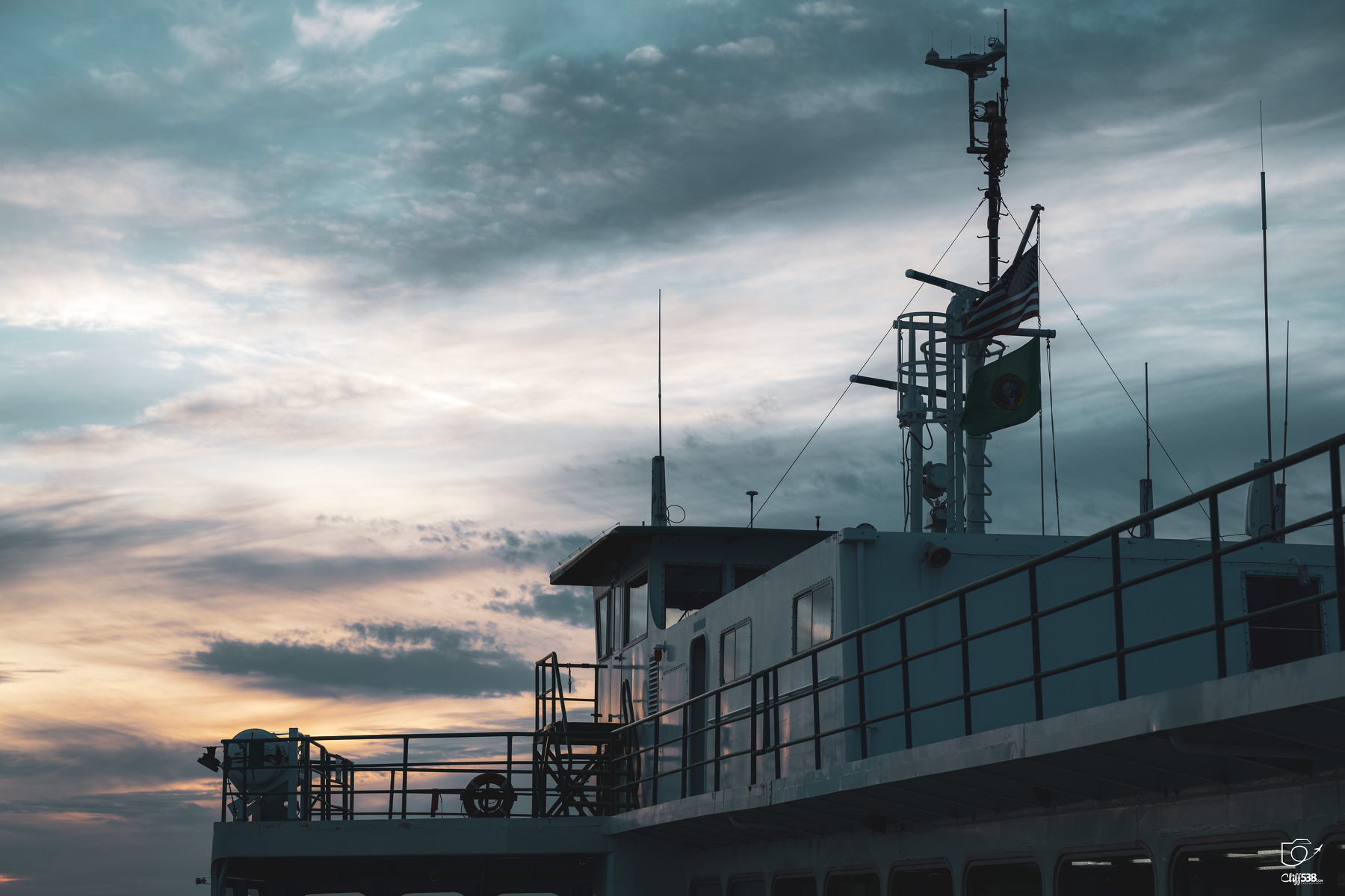 Auto-generated description: A boat or ship cabin with antennas and flags is set against a partly cloudy evening sky.