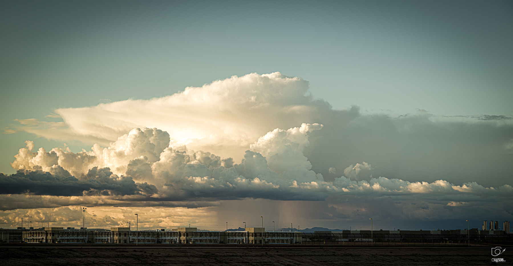 Auto-generated description: A vast sky filled with large, dramatic clouds hovers over a low-lying building complex and flat landscape.