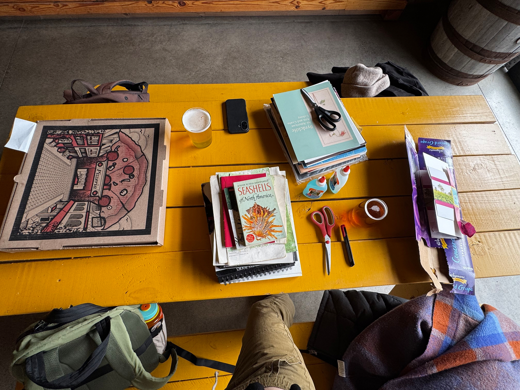 A yellow picnic table is covered with books, a pizza box, scissors, a drink, and various stationery items.