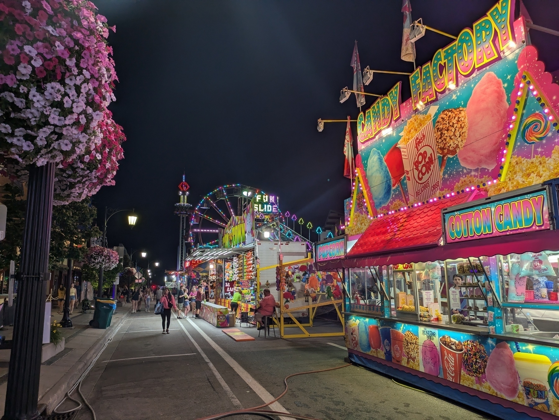 A vibrant carnival scene at night features colorful food stalls, a ferris wheel, and bustling visitors.