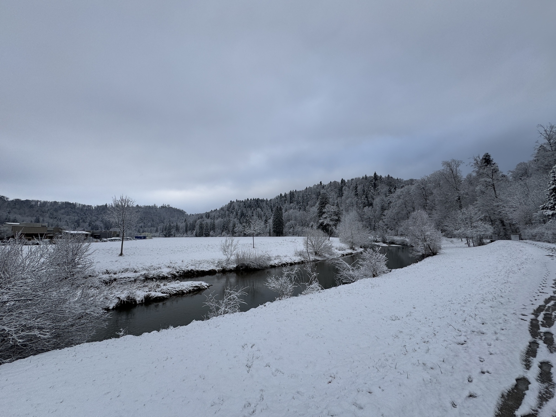 A snowy landscape features a river, snow-covered trees, open fields, and a cloudy sky.