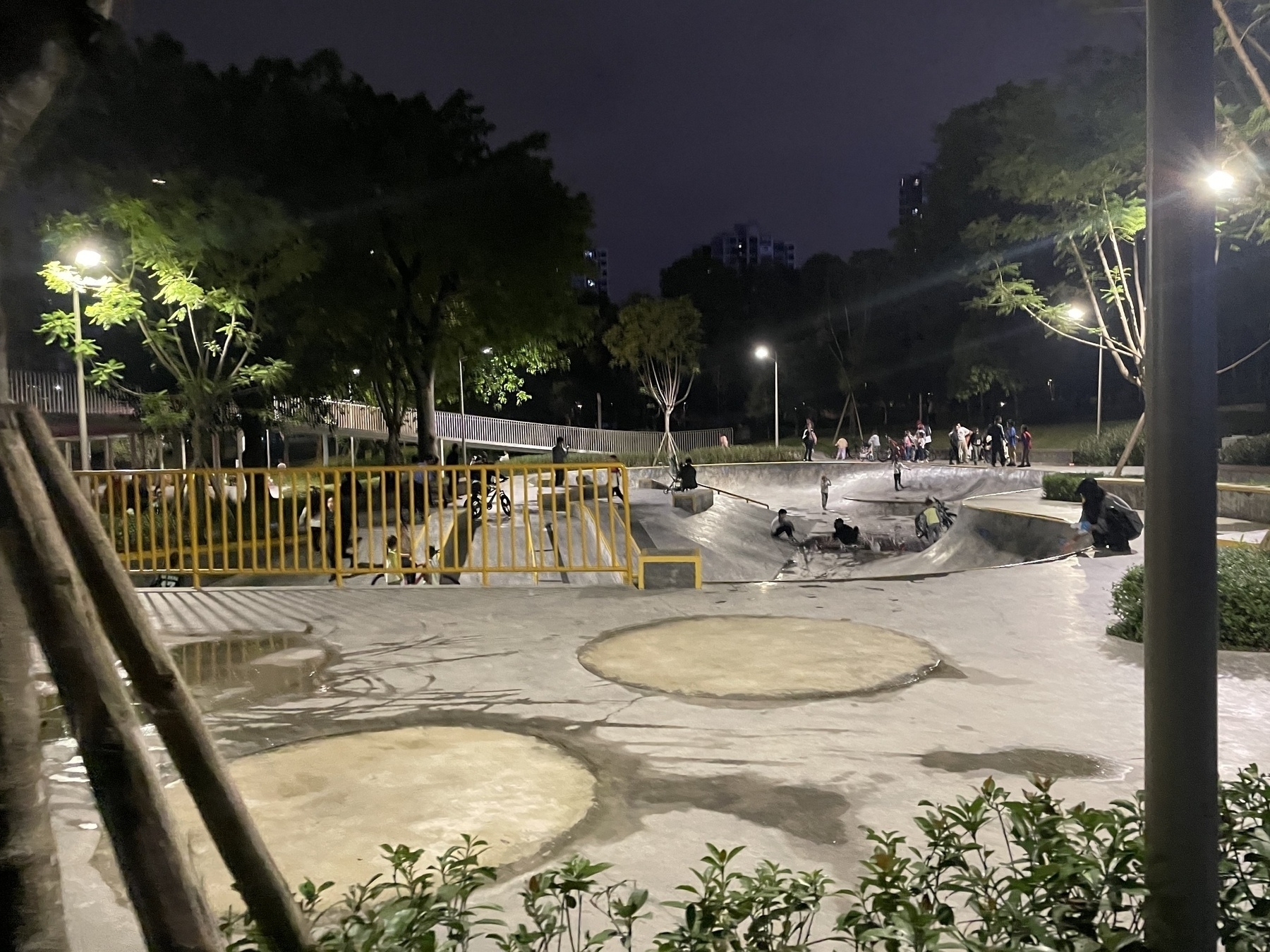 A skate park is bustling with people under bright lights at night, surrounded by trees and greenery.