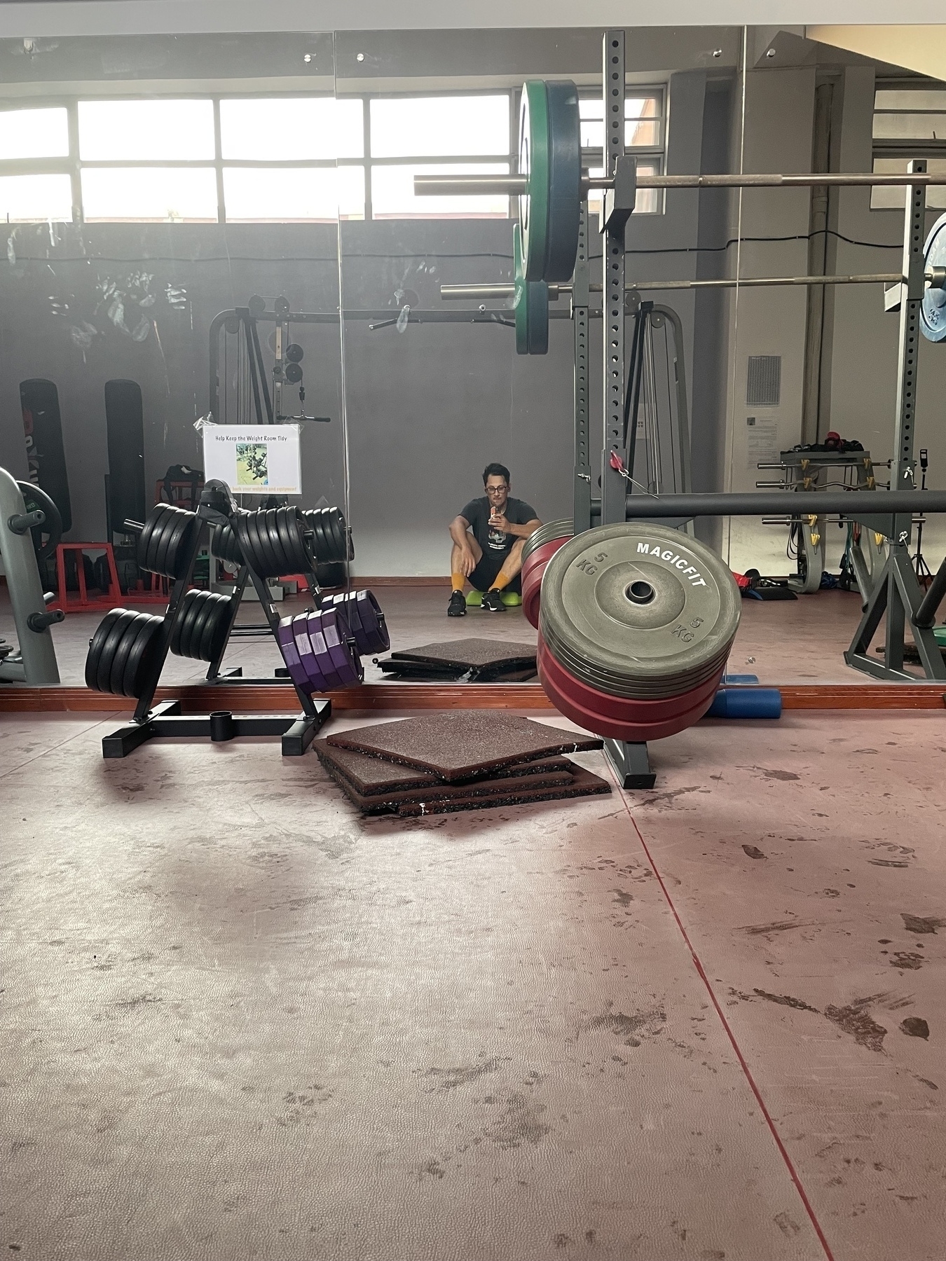 A gym setting with various weights and equipment, featuring a person sitting in the background.