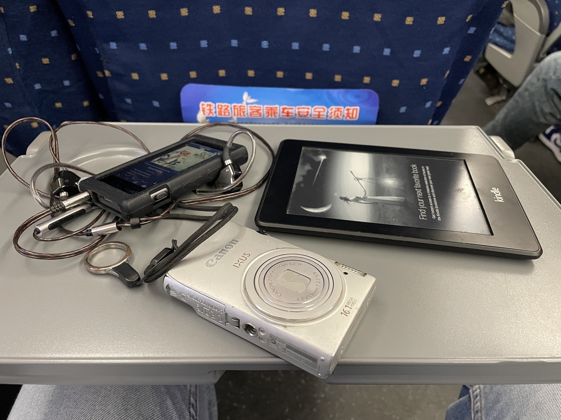 photo of a Sony mp3 player, old Canon Ixus camera and a Kindle on a fold down train table