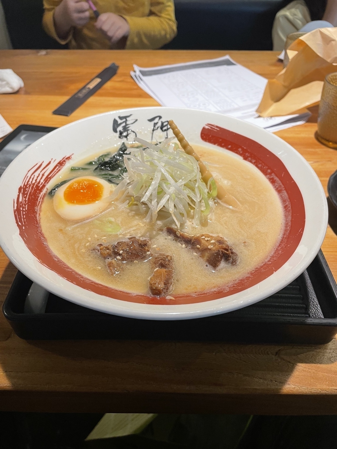 A bowl of ramen with sliced meat, bean sprouts, a halved egg, and broth is on a table with papers and a person in the background.