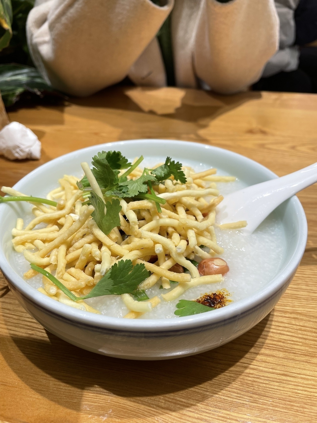 A bowl of porridge topped with crispy noodles, cilantro, and peanuts is set on a wooden table.