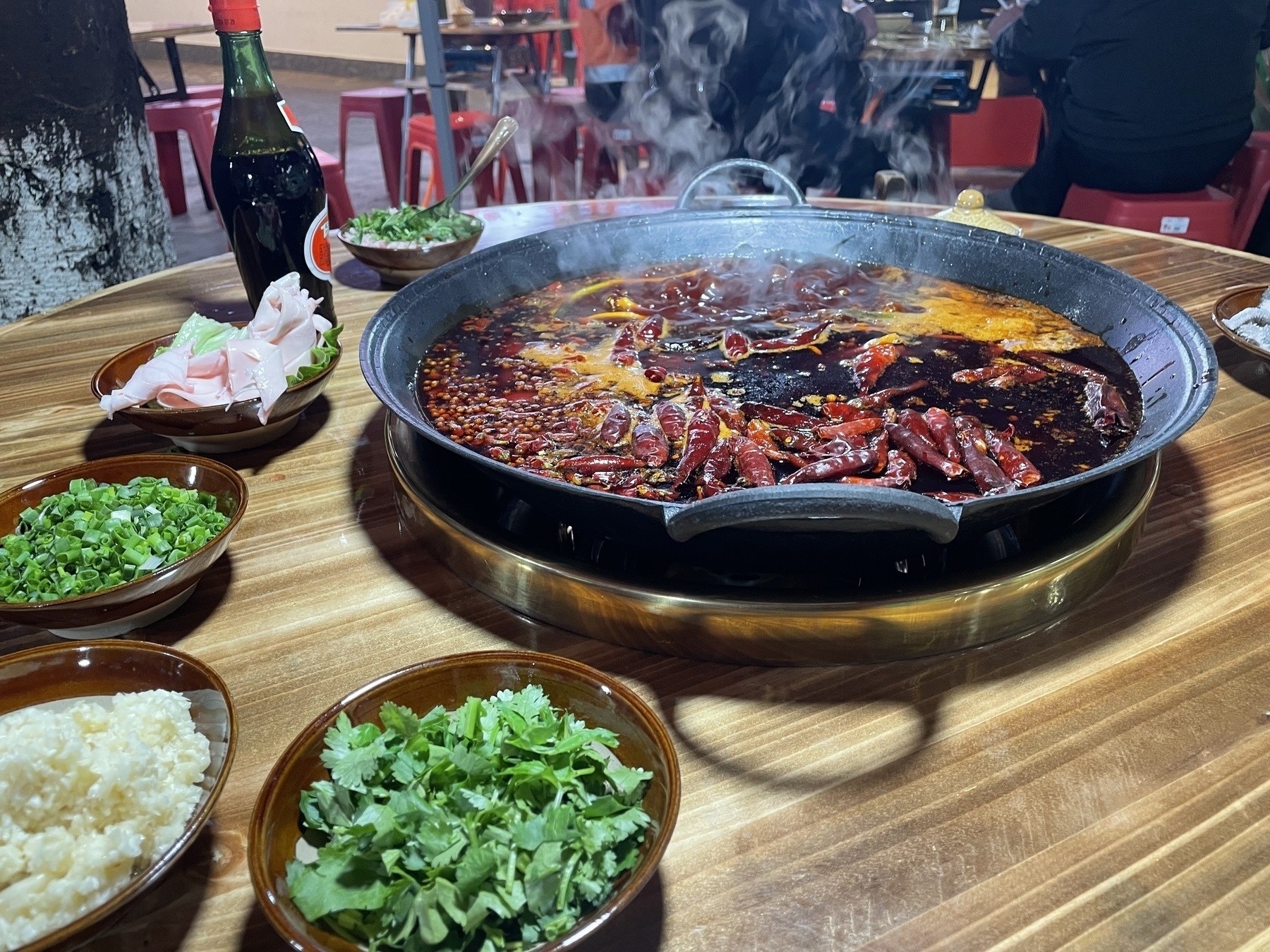 A steaming pot of hot pot with various fresh ingredients and garnishes is arranged on a wooden table.