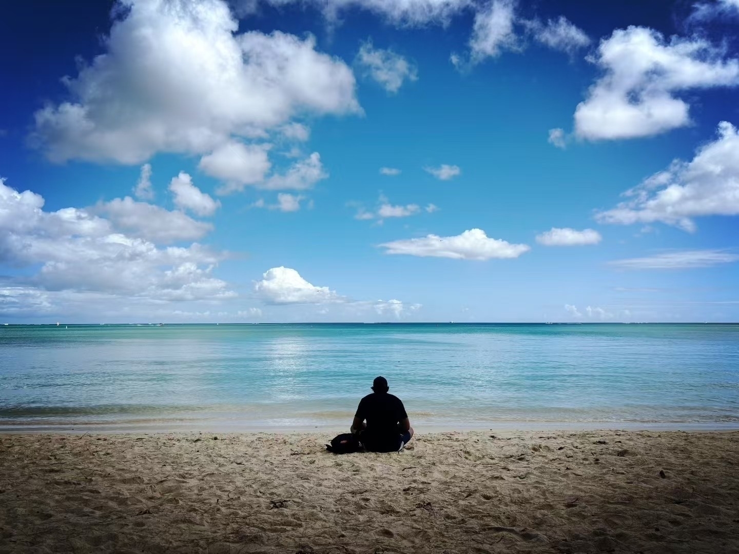 A person sits on a sandy beach facing a calm, expansive ocean under a partly cloudy sky.