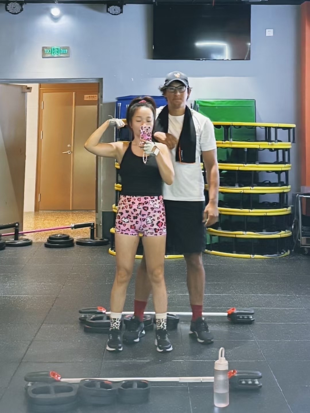 Two people are taking a gym selfie in front of a mirror, surrounded by fitness equipment.