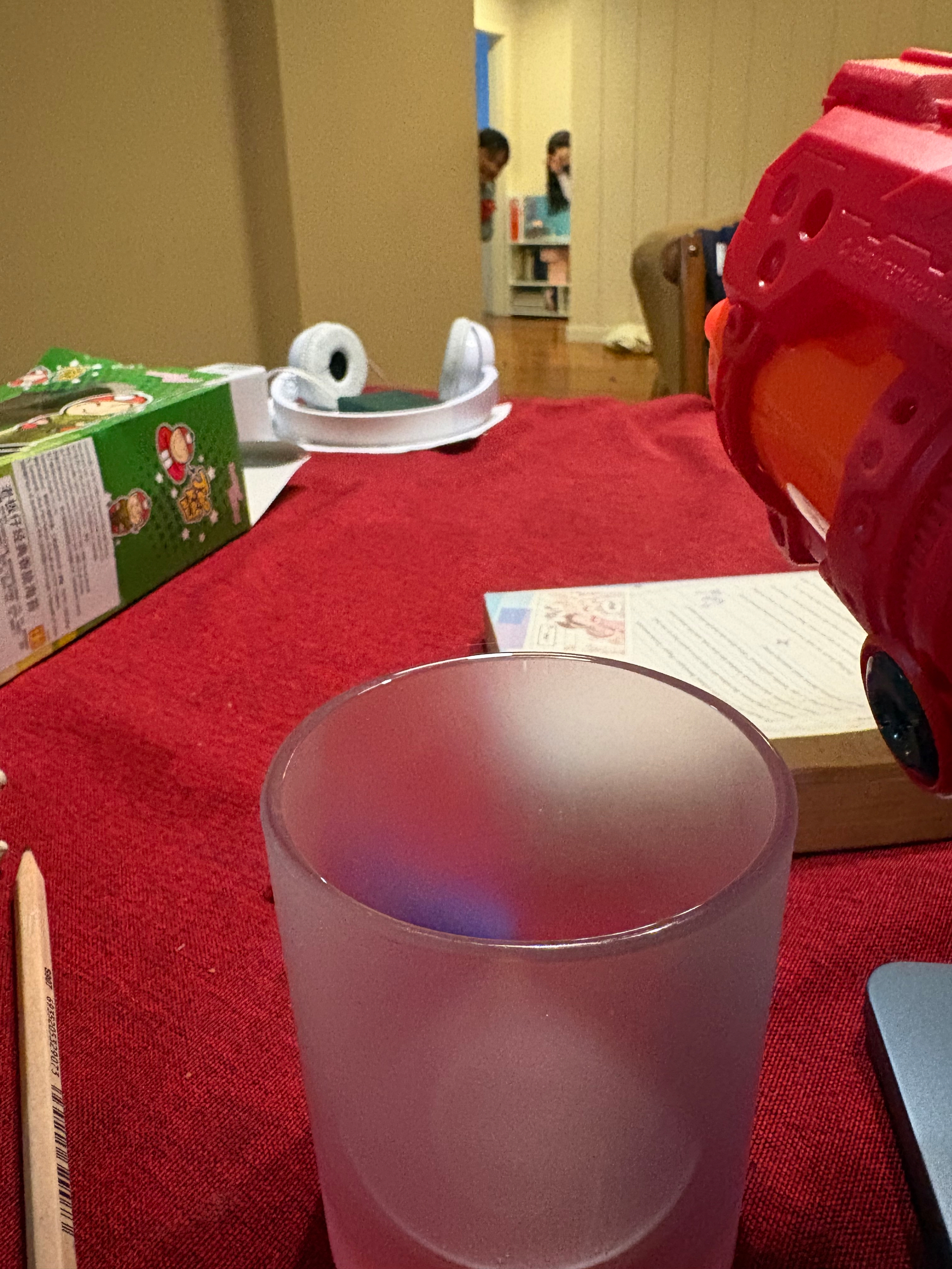 A table with a glass, a pencil, and a toy, with people and a box in the background.