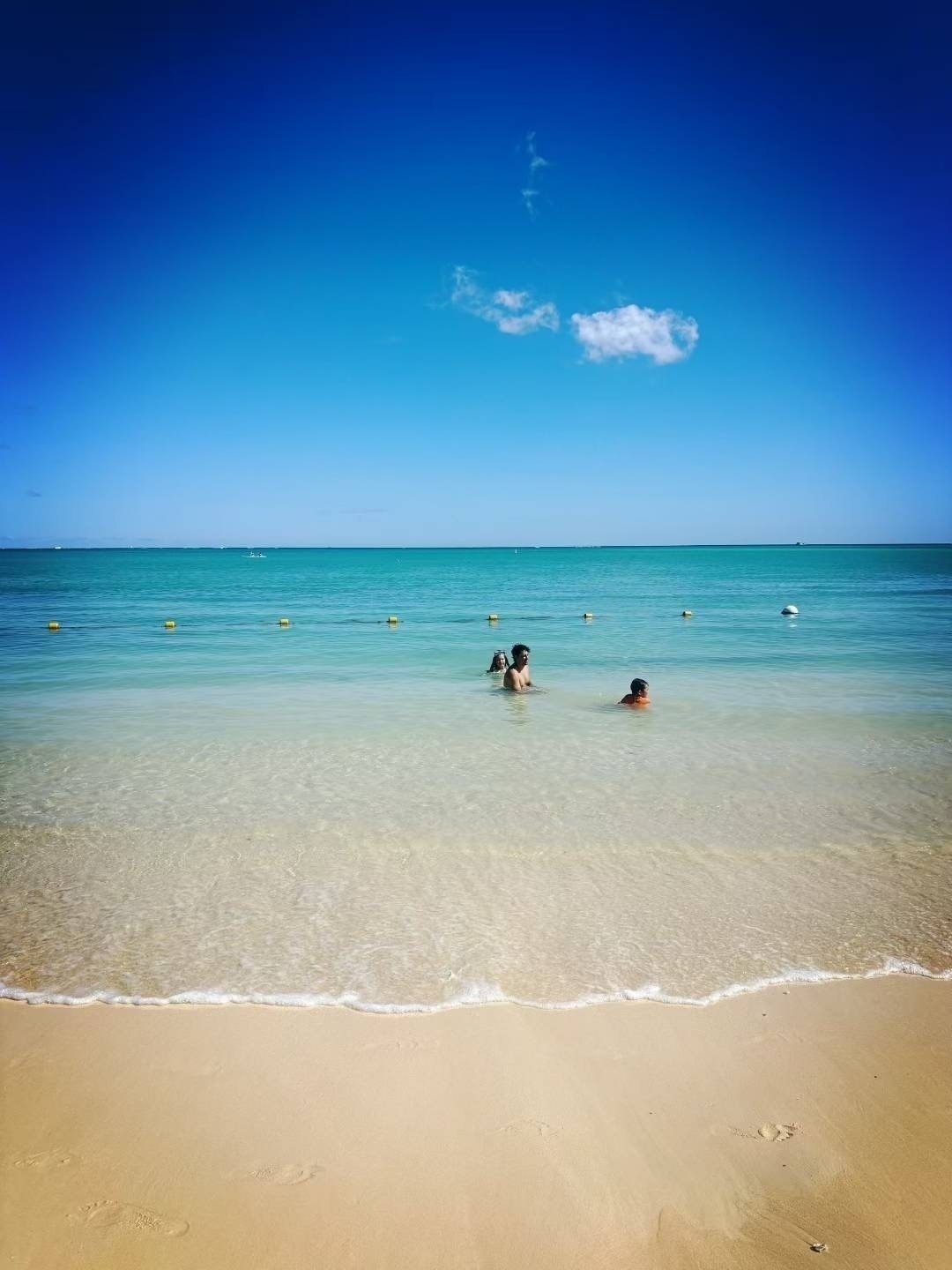 A few people are swimming in the clear, shallow waters of a beach under a blue sky with a single cloud.