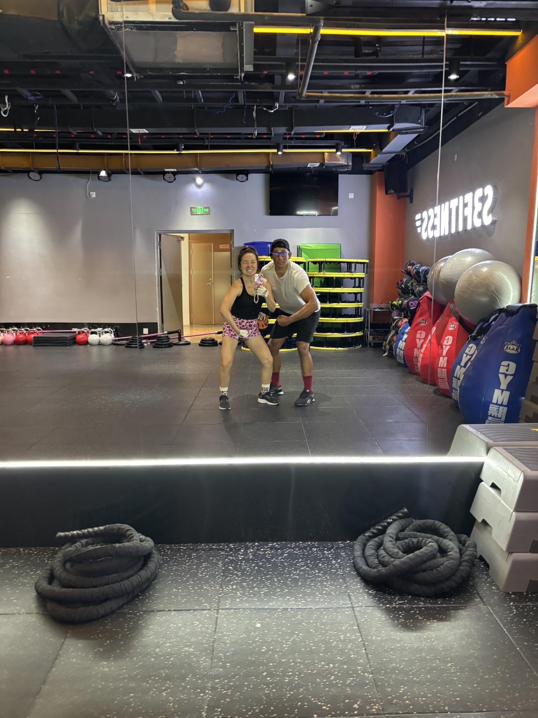 Two people smiling and posing for a selfie in a gym with boxing equipment and ropes visible.