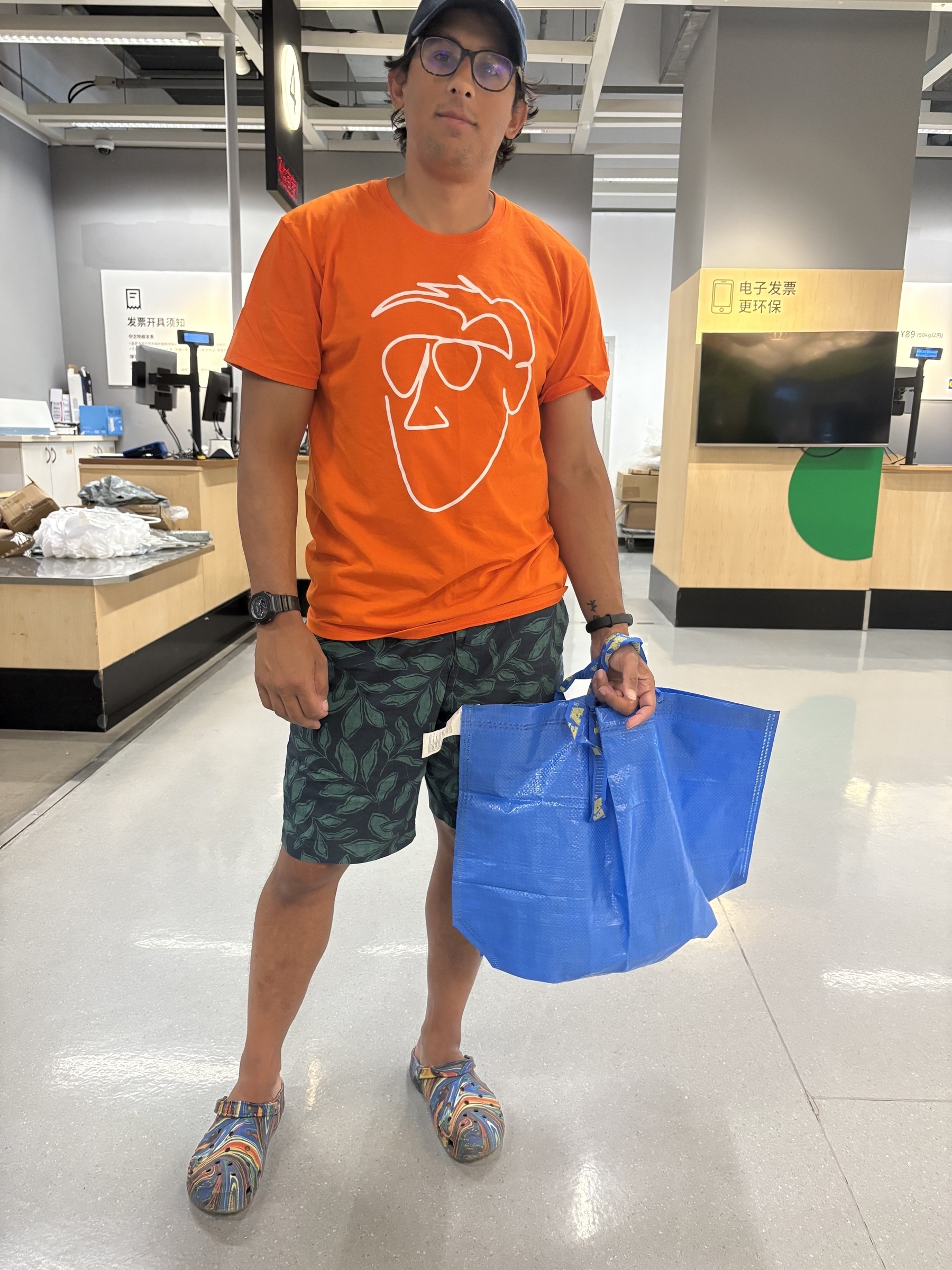A person in casual attire, including an orange t-shirt and patterned shorts, is standing indoors holding blue shopping bags.