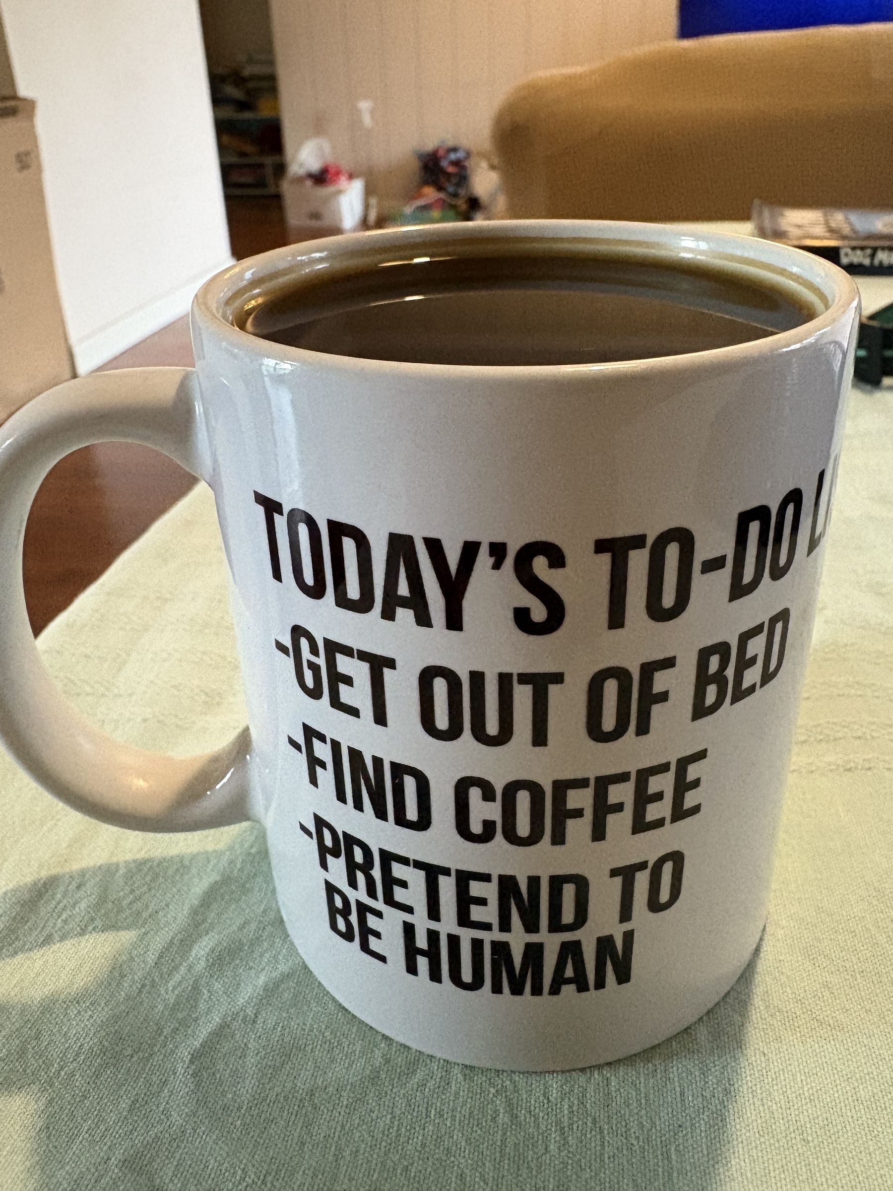 A white mug with humorous text about a to-do list, featuring tasks like getting out of bed, finding coffee, and pretending to be human, is filled with coffee.