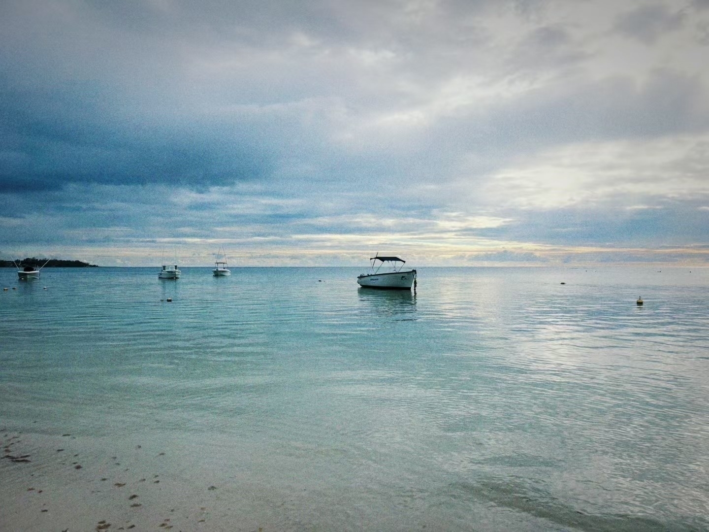 A serene beach scene features calm waters with several boats floating, under a cloudy sky.