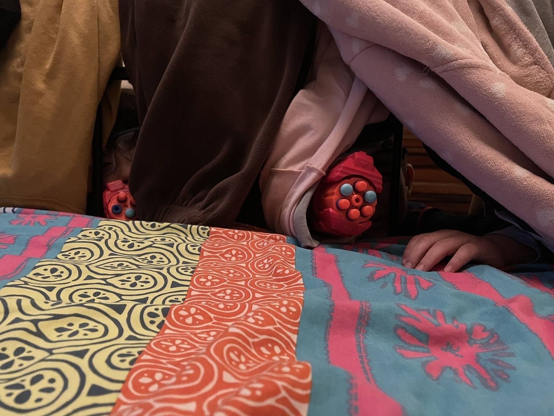 Children are partially hidden under a pile of colorful blankets on a patterned bed.