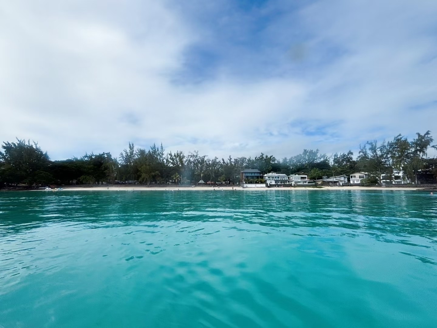 A serene turquoise seascape stretches towards a distant tree-lined shoreline under a partly cloudy sky.