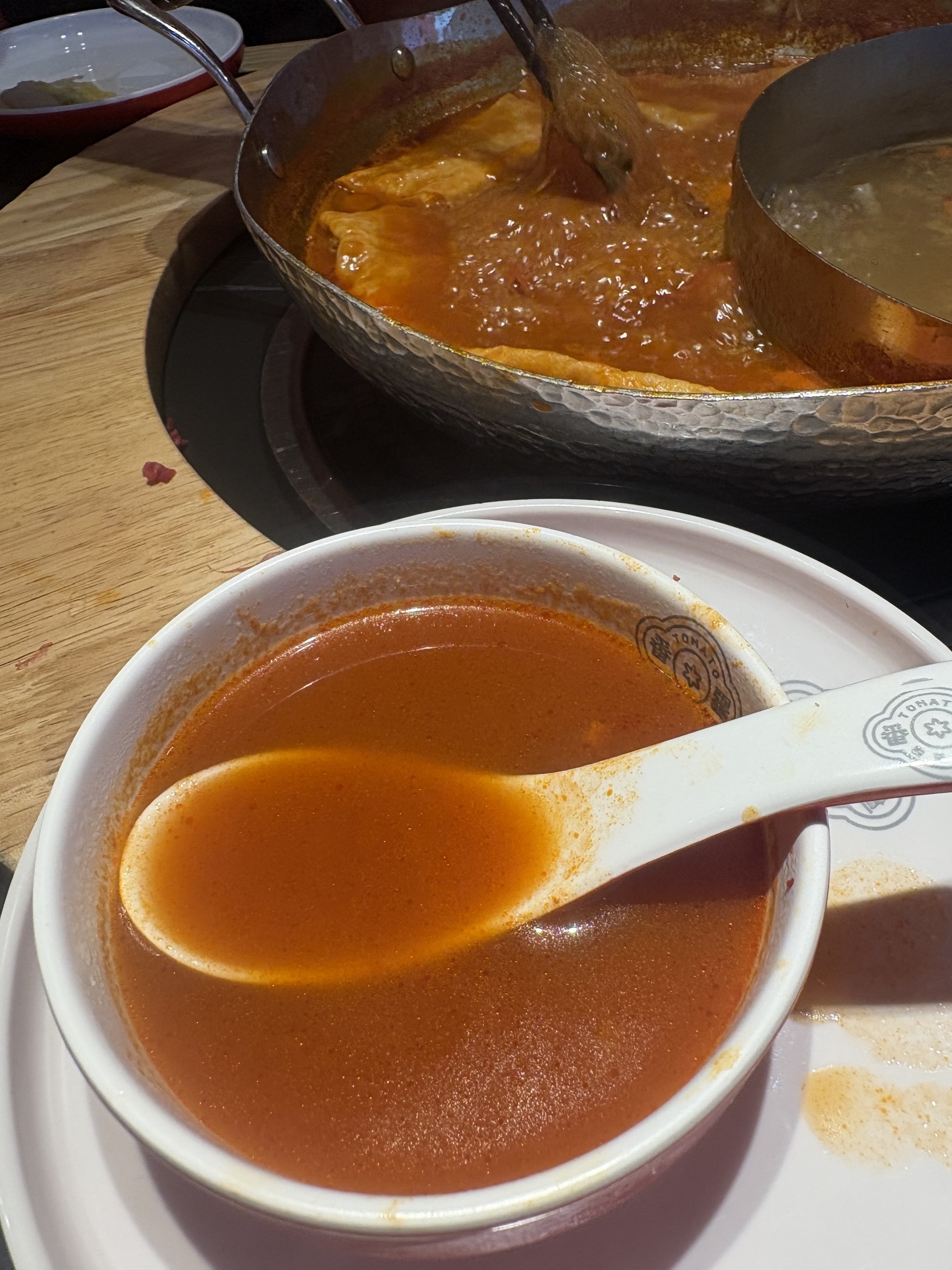 A bowl of rich, red soup sits on a plate with a white spoon, in front of a larger pot of simmering broth.