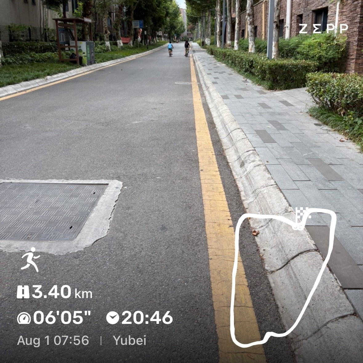 A quiet street with a yellow line marking the edge, bordered by a sidewalk and greenery, where two cyclists are visible in the distance.