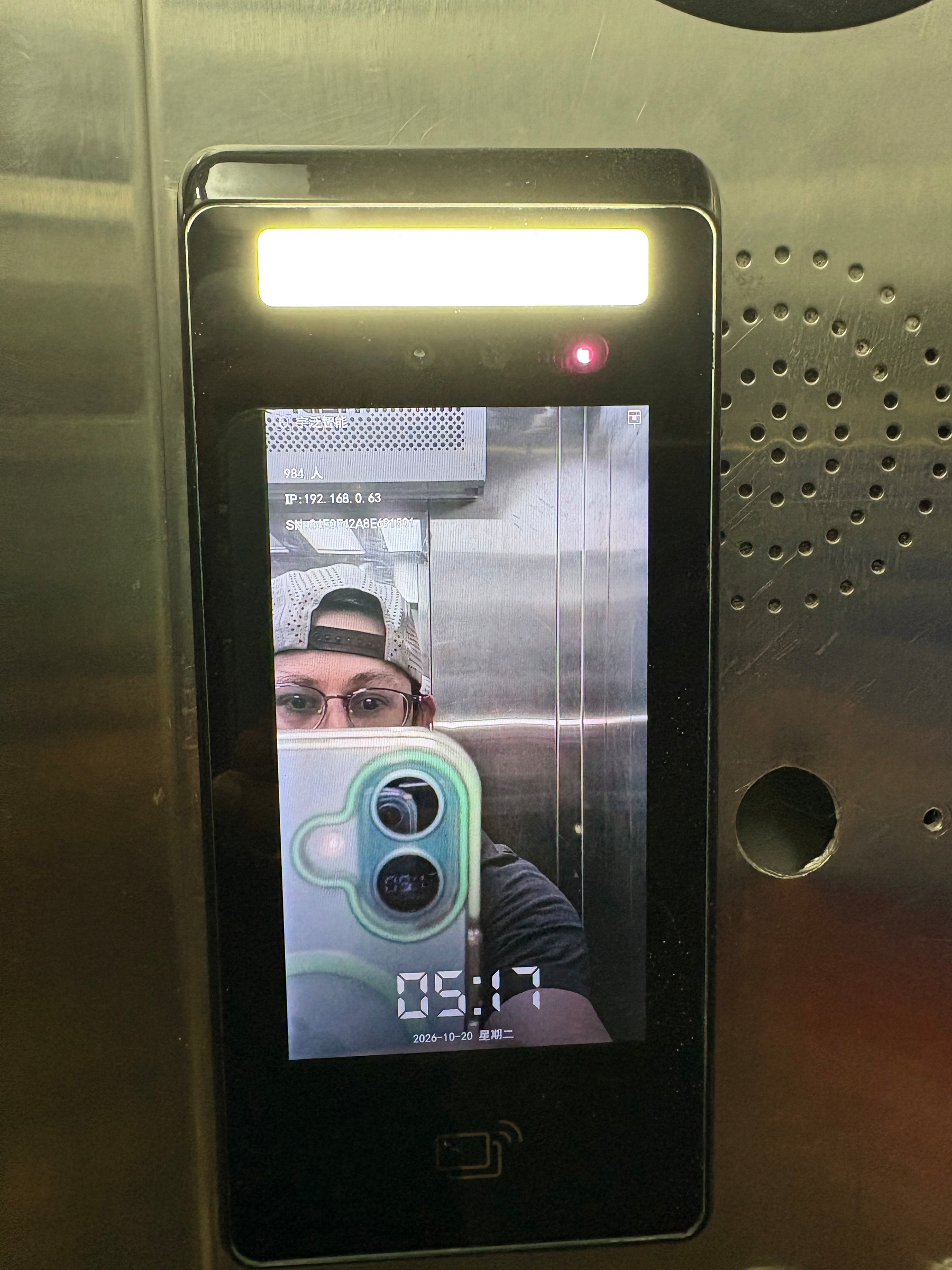 A person wearing glasses and a backwards cap is taking a selfie, which is displayed on a digital screen with a timestamp of 05:17.