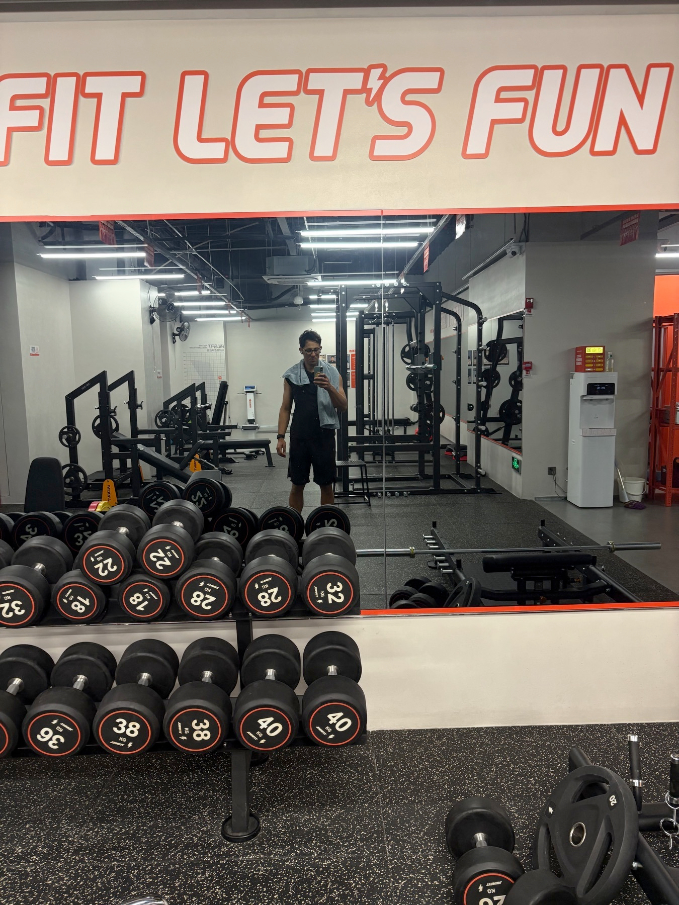 A person is taking a mirror selfie in a gym with various weights and equipment around.