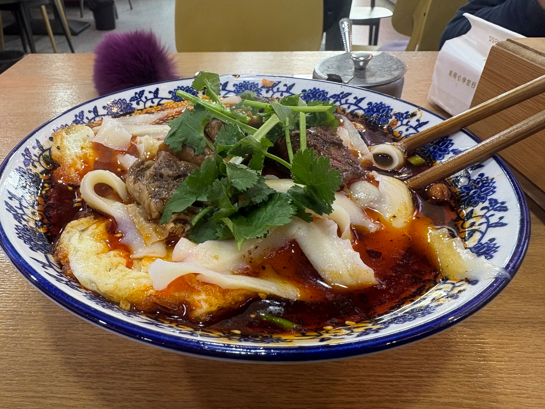 A bowl of spicy noodle soup topped with herbs and meat is served on a decorated blue and white dish.