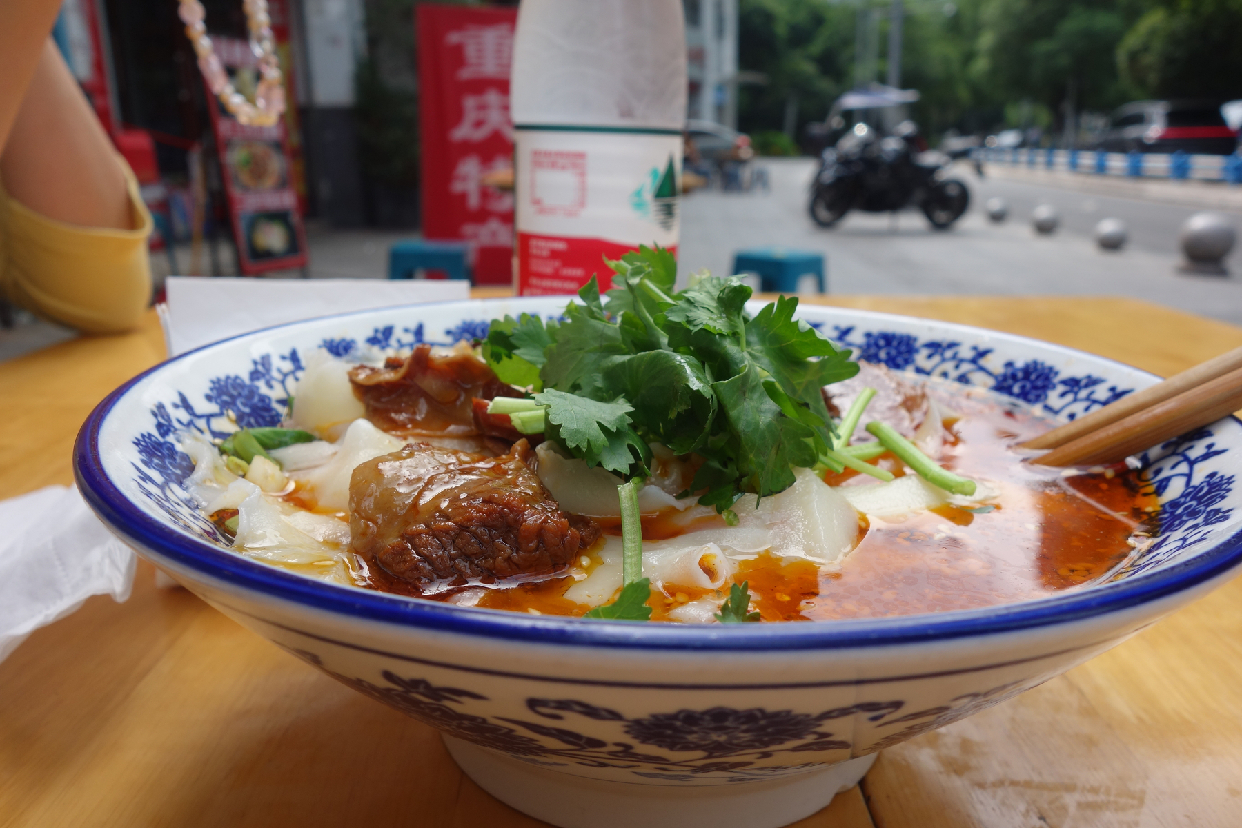 A bowl of spicy, beef noodles topped with fresh coriander is placed on a wooden table outdoors.