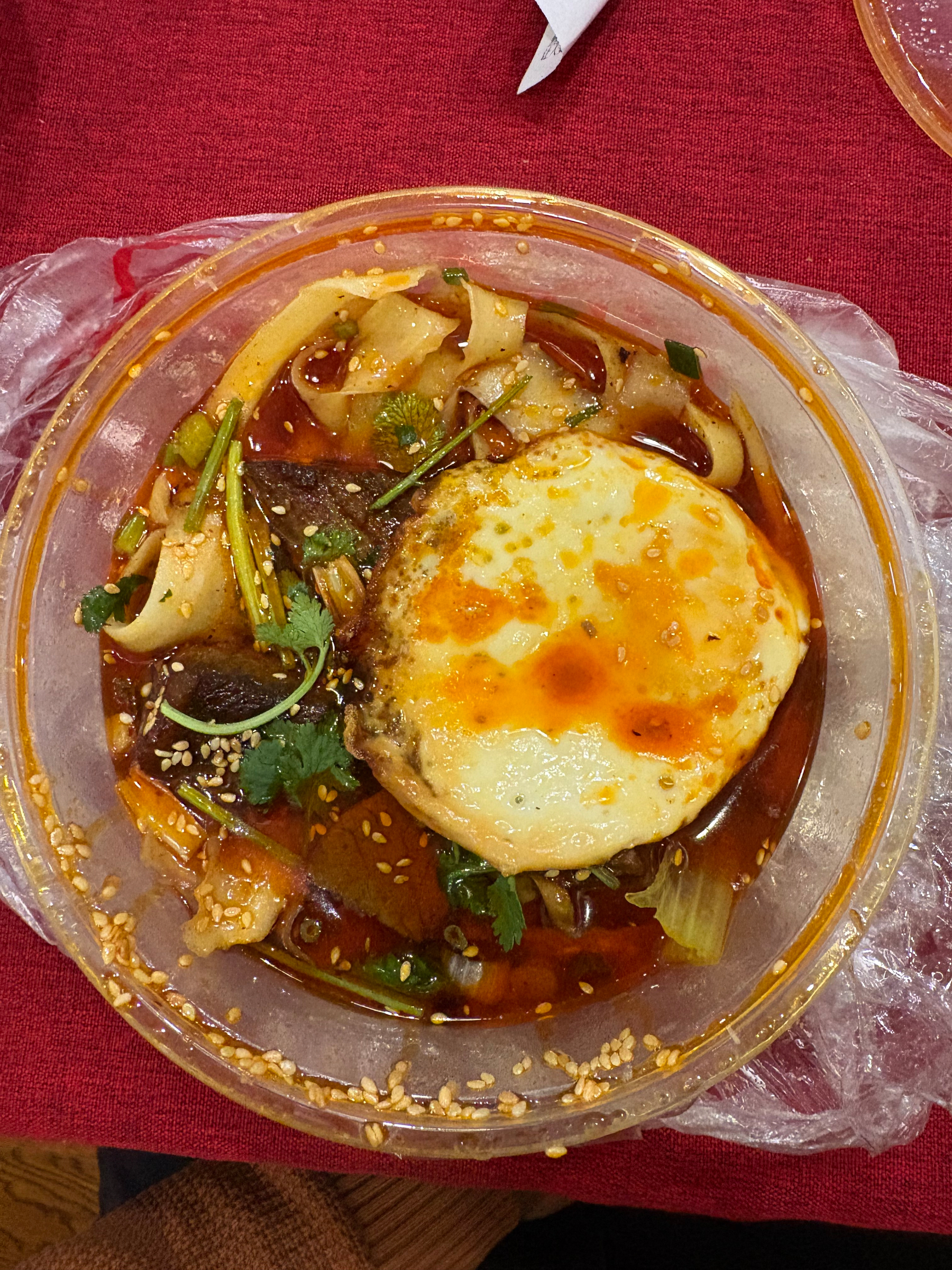A bowl of spicy noodle soup is topped with a fried egg, garnished with coriander and sesame seeds.