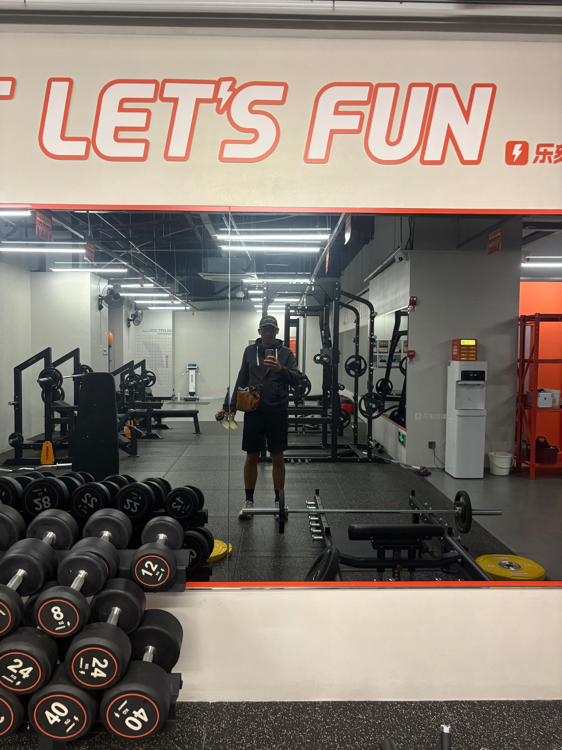 A person is taking a selfie in a gym with various weights, exercise machines, and a large wall mirror.