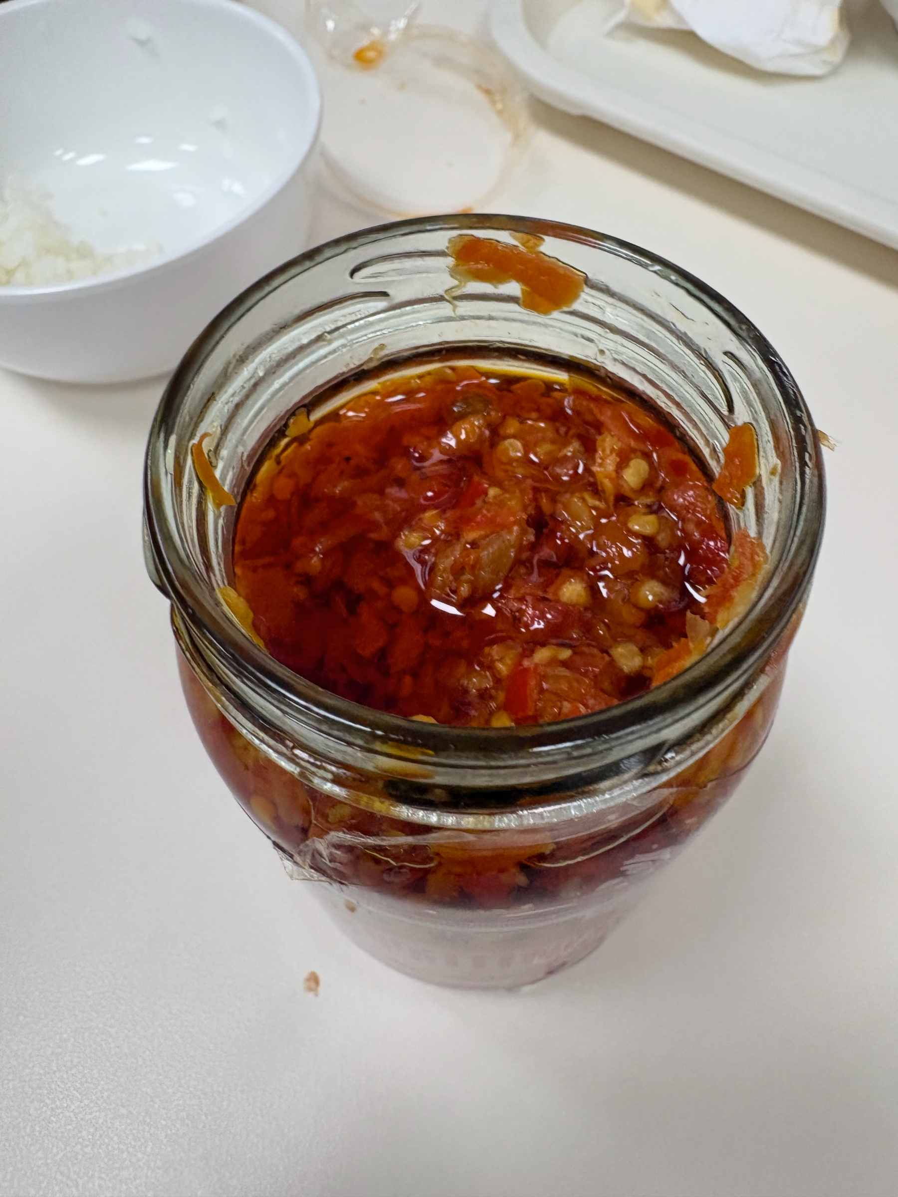 A jar filled with chili oil sits on a table, accompanied by a bowl and a tray in the background.