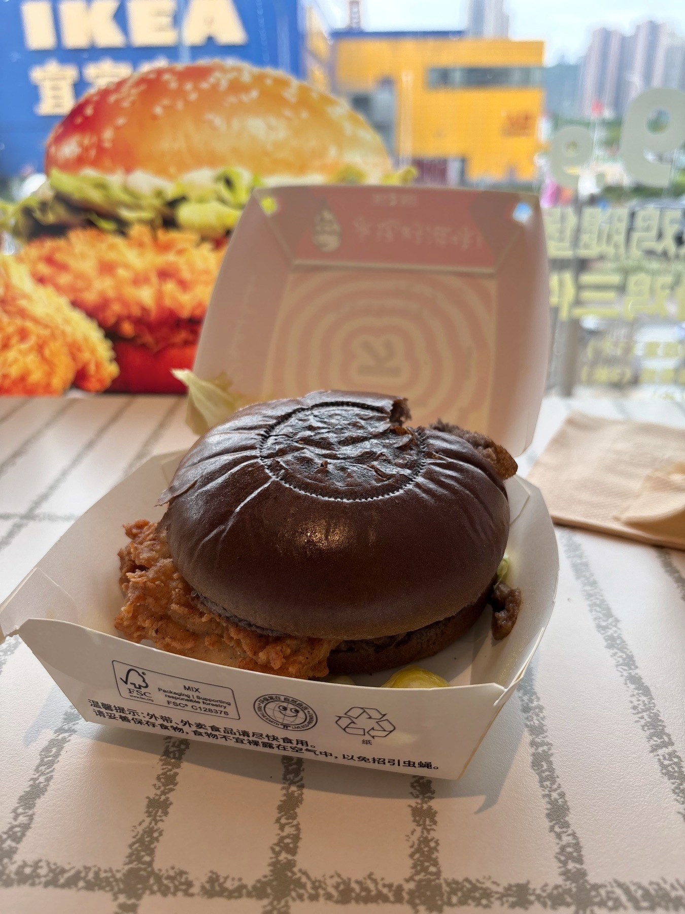 A crispy fried chicken sandwich with a dark bun is served in a white takeout container on a table with an IKEA-themed advertisement in the background.