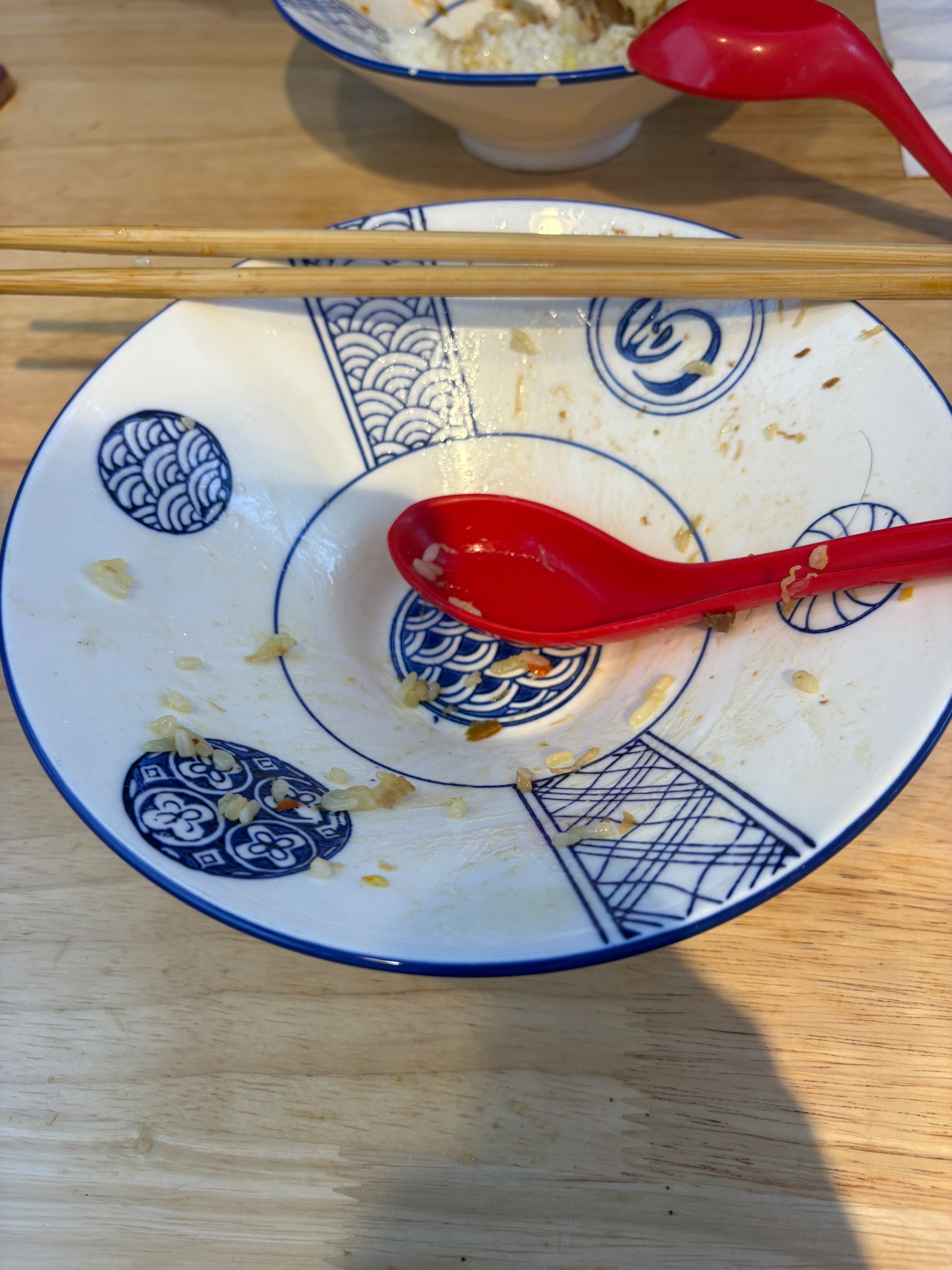 A patterned bowl with a red spoon and chopsticks rests on a wooden table, containing remnants of food.