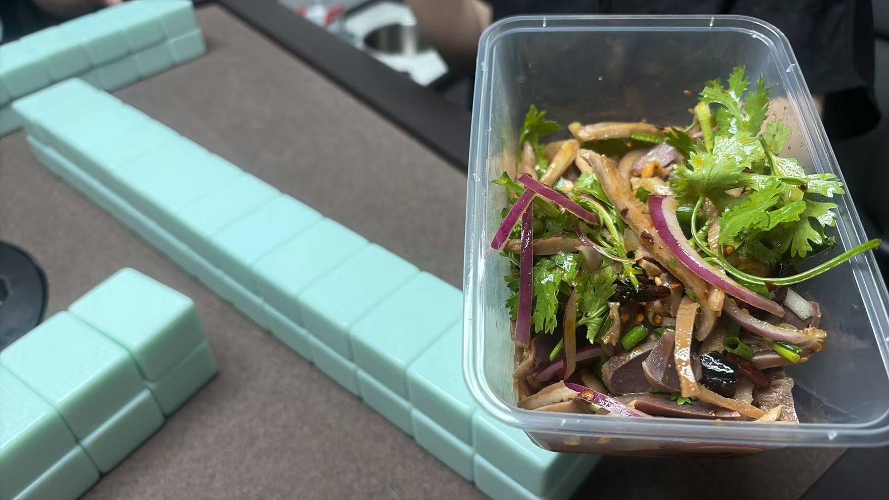 A container of mixed salad with herbs and sliced vegetables is placed next to a row of light blue majiang tiles.