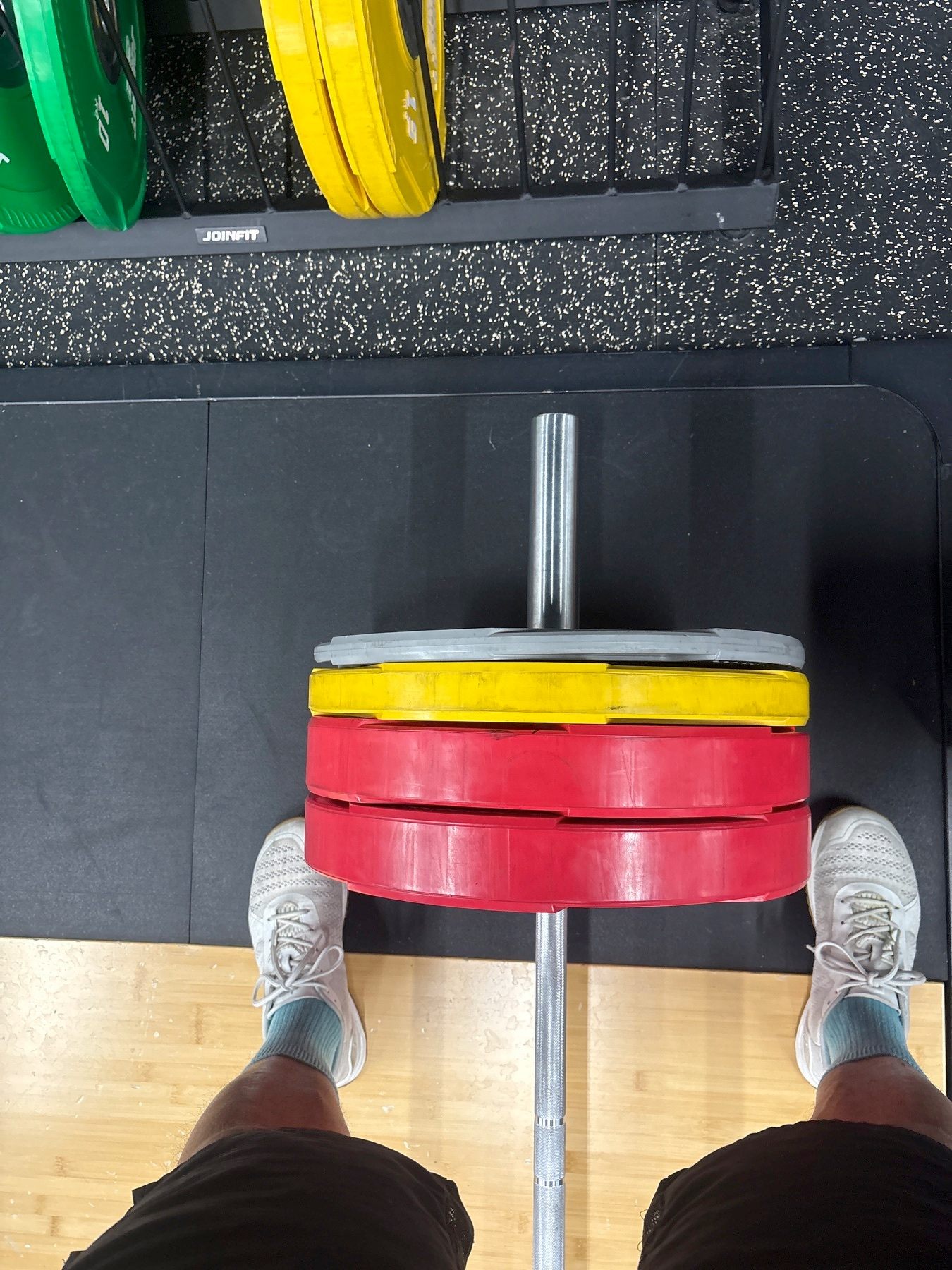 A person wearing white sneakers stands in front of a barbell with red, yellow, and gray weight plates, placed on a gym floor next to a weight rack.