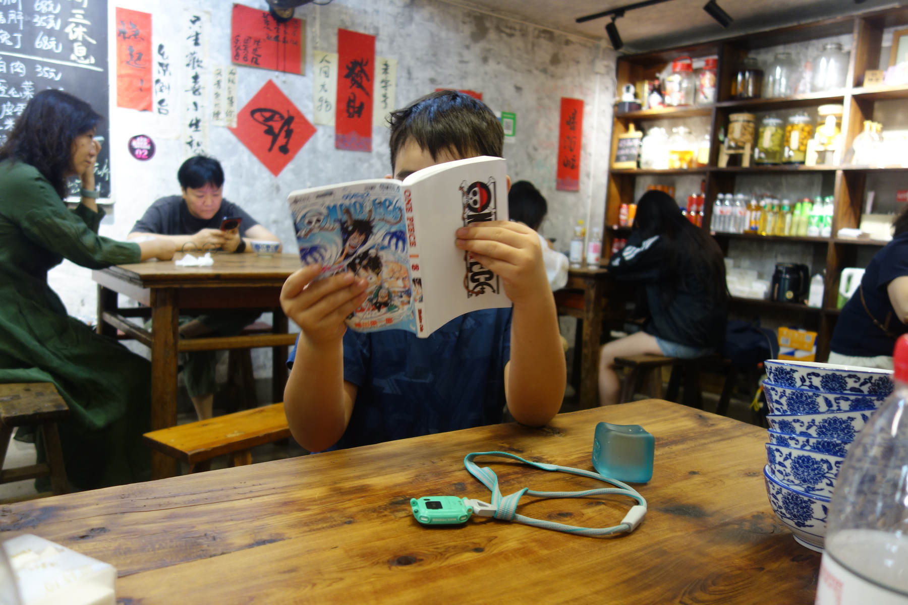 A child is sitting at a wooden table in a cozy room, reading a manga book while other people are seated and engaged with their phones or conversations.
