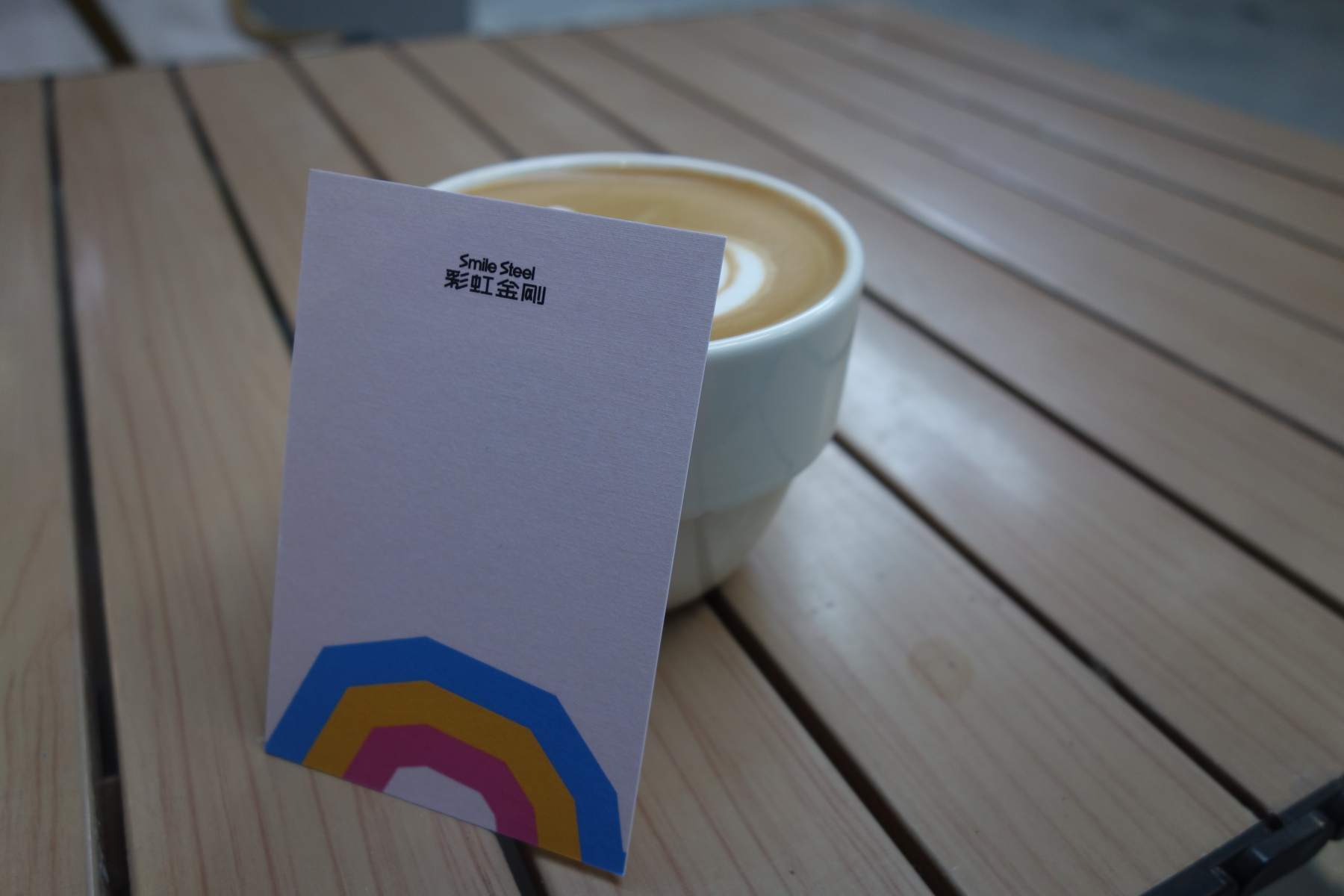 A cup of coffee with latte art is placed on a wooden table, accompanied by a card featuring colorful geometric shapes and text in both English and Chinese.