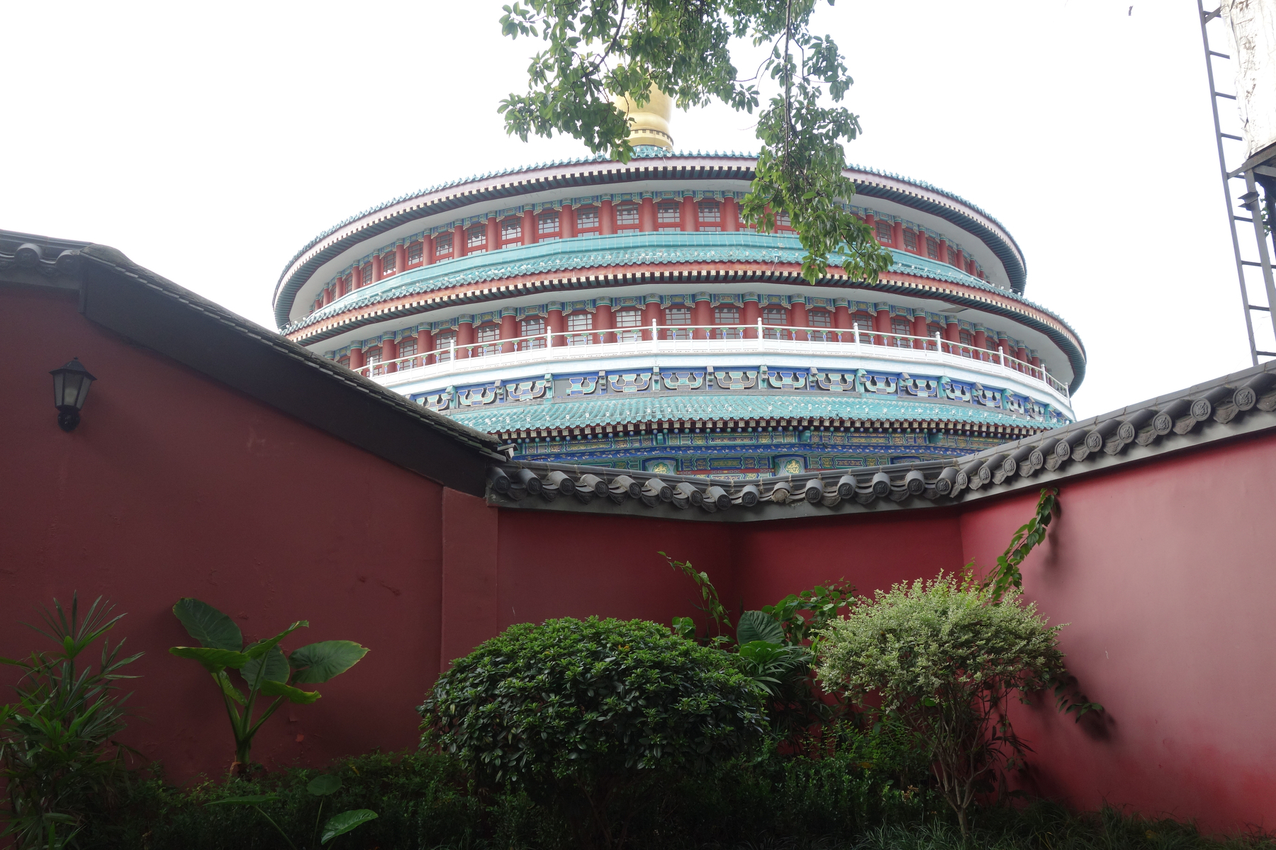 A colorful, traditional circular building with intricate architectural details is partially visible behind a red wall and green foliage.