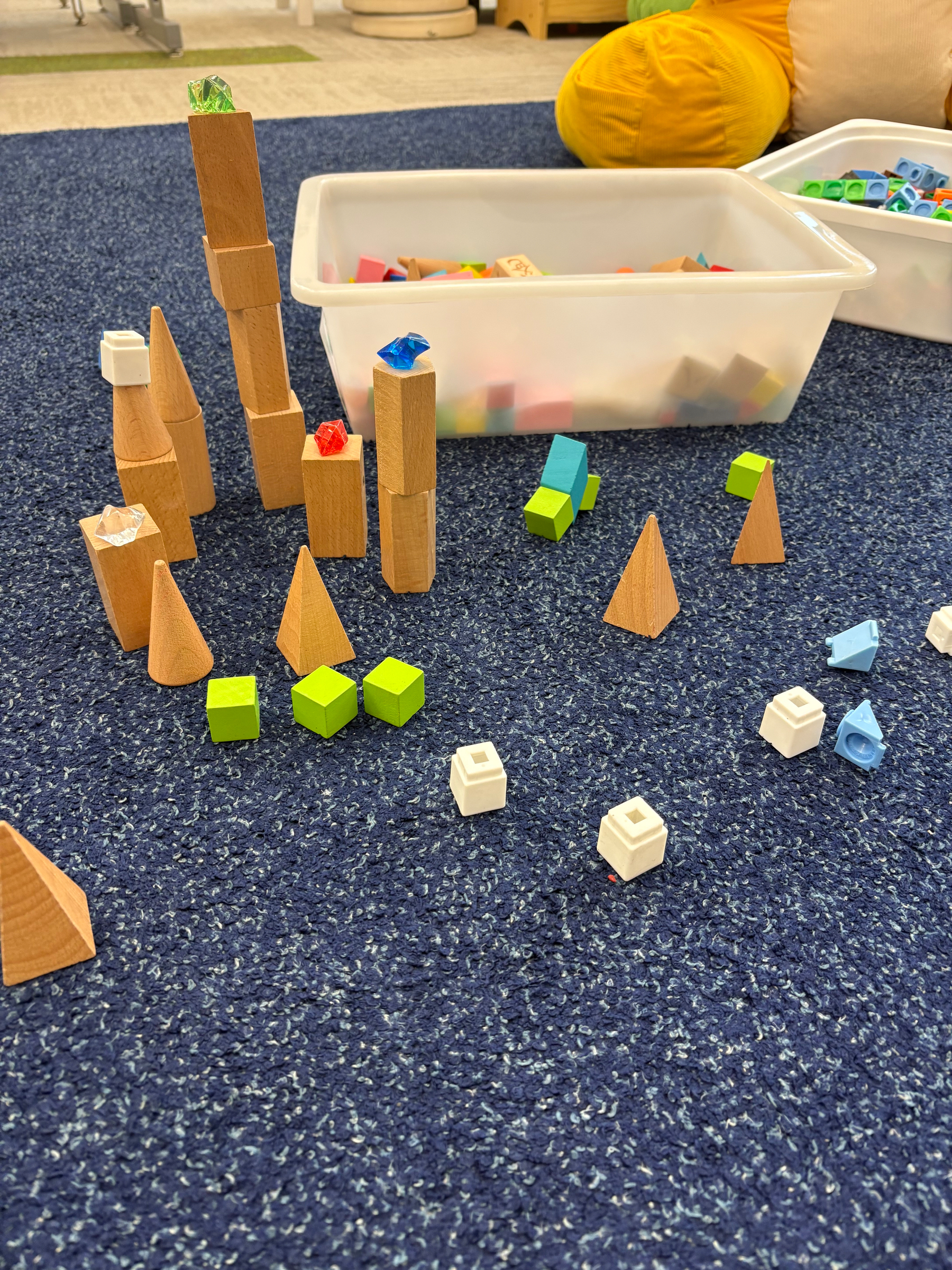 Various wooden blocks and colorful plastic pieces are arranged on a blue carpet next to white plastic bins filled with more building toys.