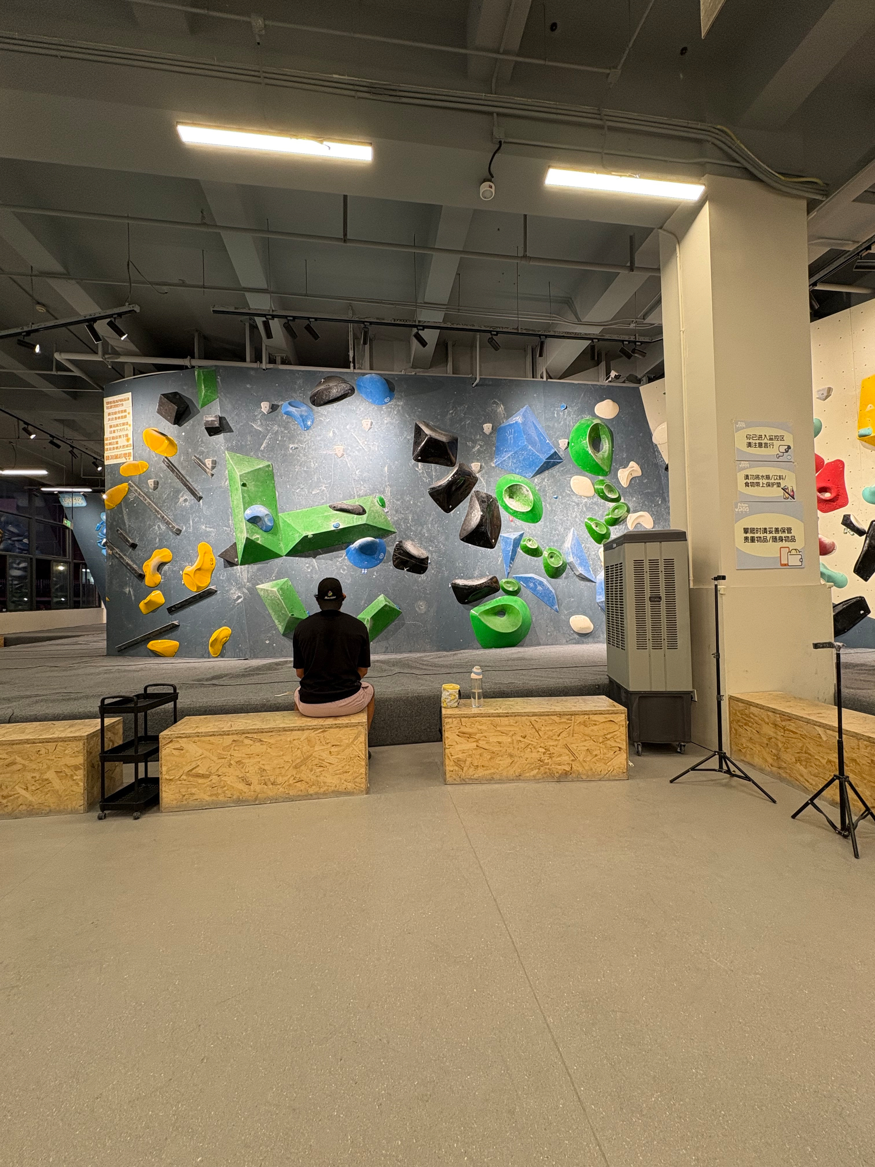 A person wearing a black shirt and cap is sitting on a wooden bench facing an indoor climbing wall with various colorful holds.