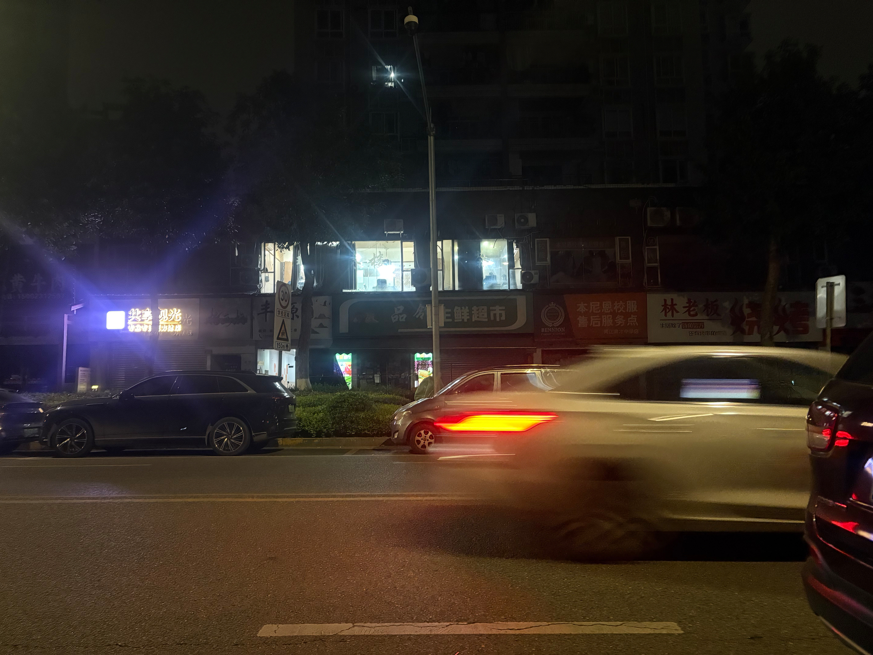A nighttime street scene features a blurry motion of a white car driving past parked vehicles and illuminated shop signs.