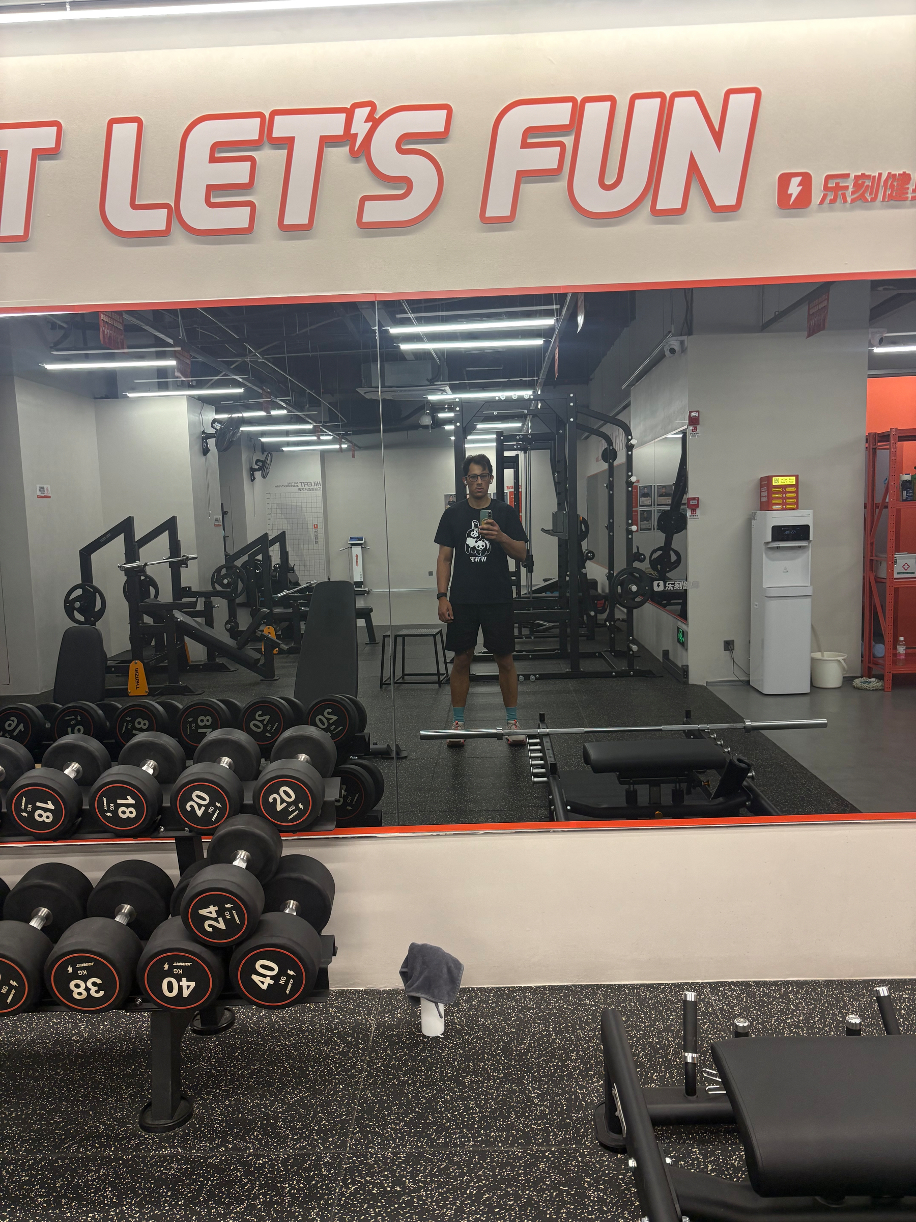 A person is taking a mirror selfie in a gym, surrounded by workout equipment and dumbbells.